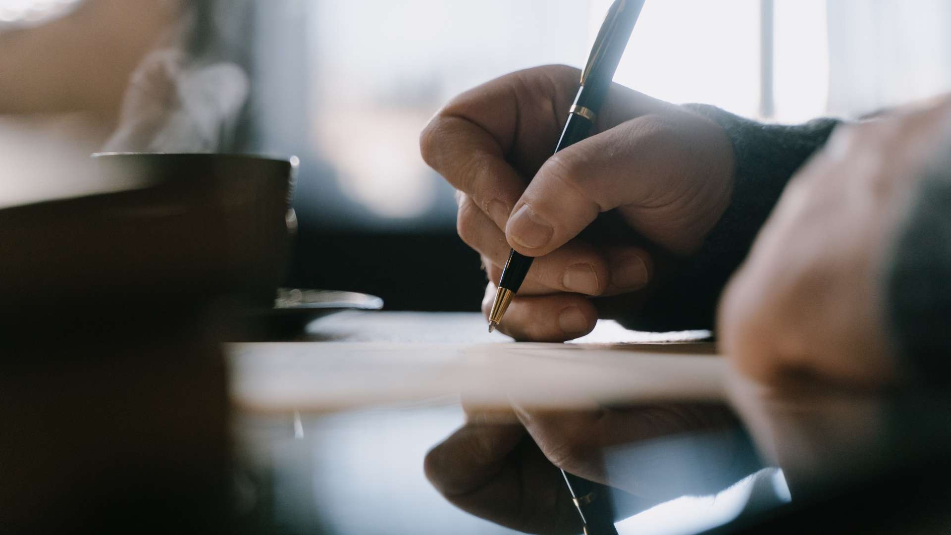A close-up of a person's hand signing an important document with a classic pen, with a steaming coffee cup nearby, signifying a serious business negotiation or agreement.