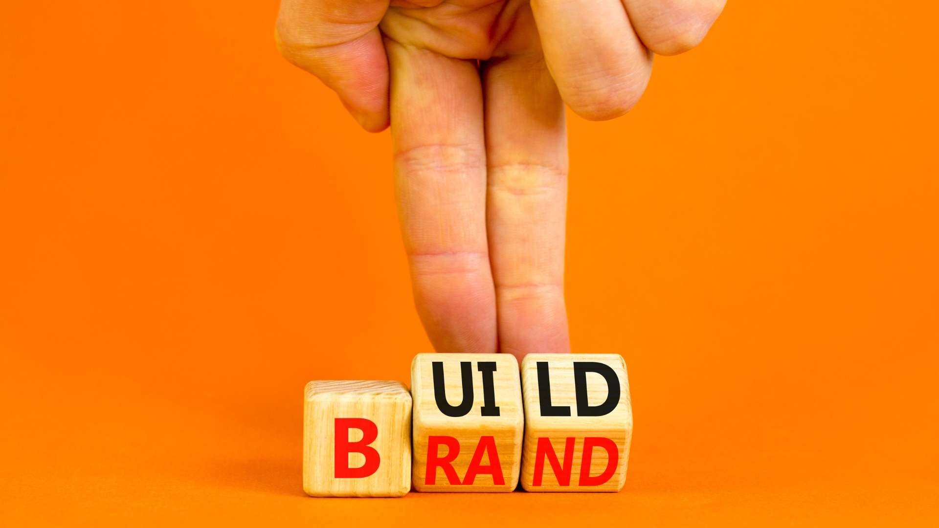 A person's fingers arrange wooden letter blocks on a bright orange surface to spell out "BUILD BRAND," representing the concept of constructing a brand identity.