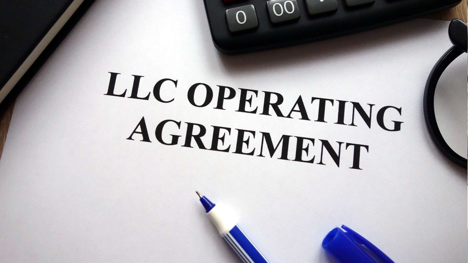 A close-up image of a document titled "LLC Operating Agreement" on a white piece of paper, accompanied by a blue pen, a pair of reading glasses, a calculator, and a black notebook on a wooden surface.