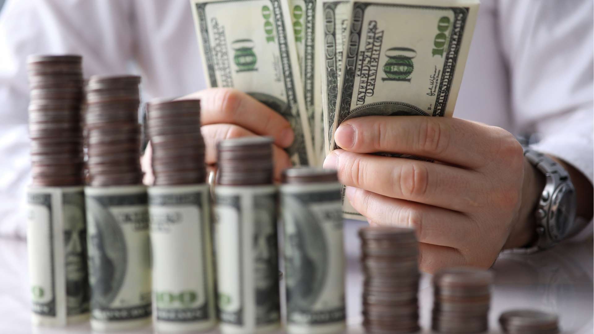 A person holding fanned-out hundred-dollar bills in their hands, with stacks of coins and rolled dollar bills on a table in the foreground.