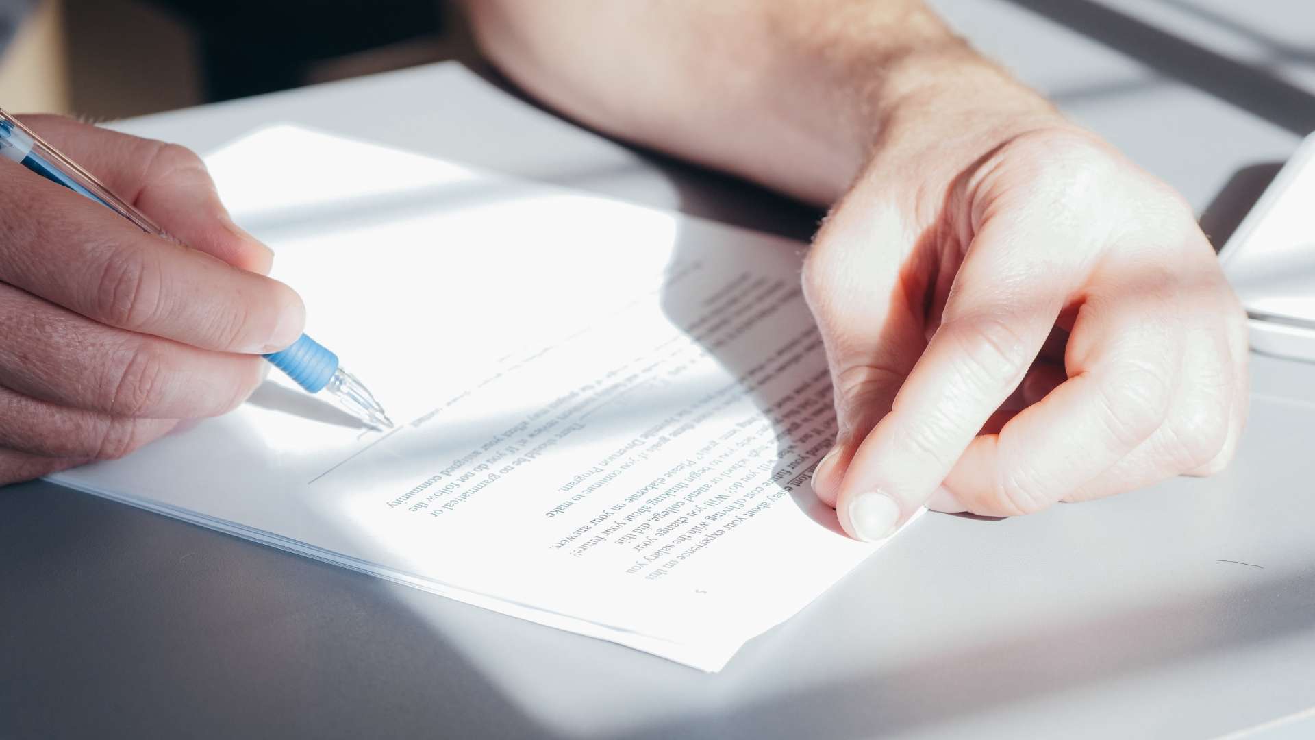 Person signing a legal document with a blue pen on a desk in natural light.