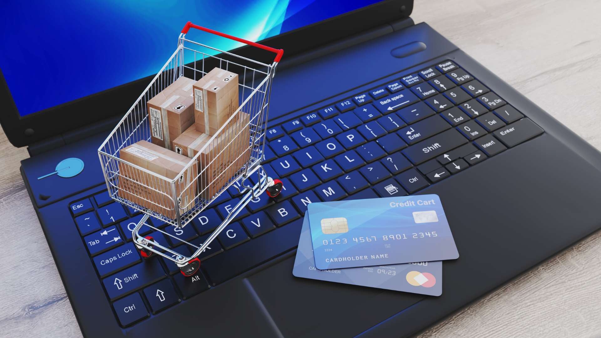 Mini shopping cart filled with packages on a laptop keyboard with credit cards, symbolizing e-commerce and online shopping.