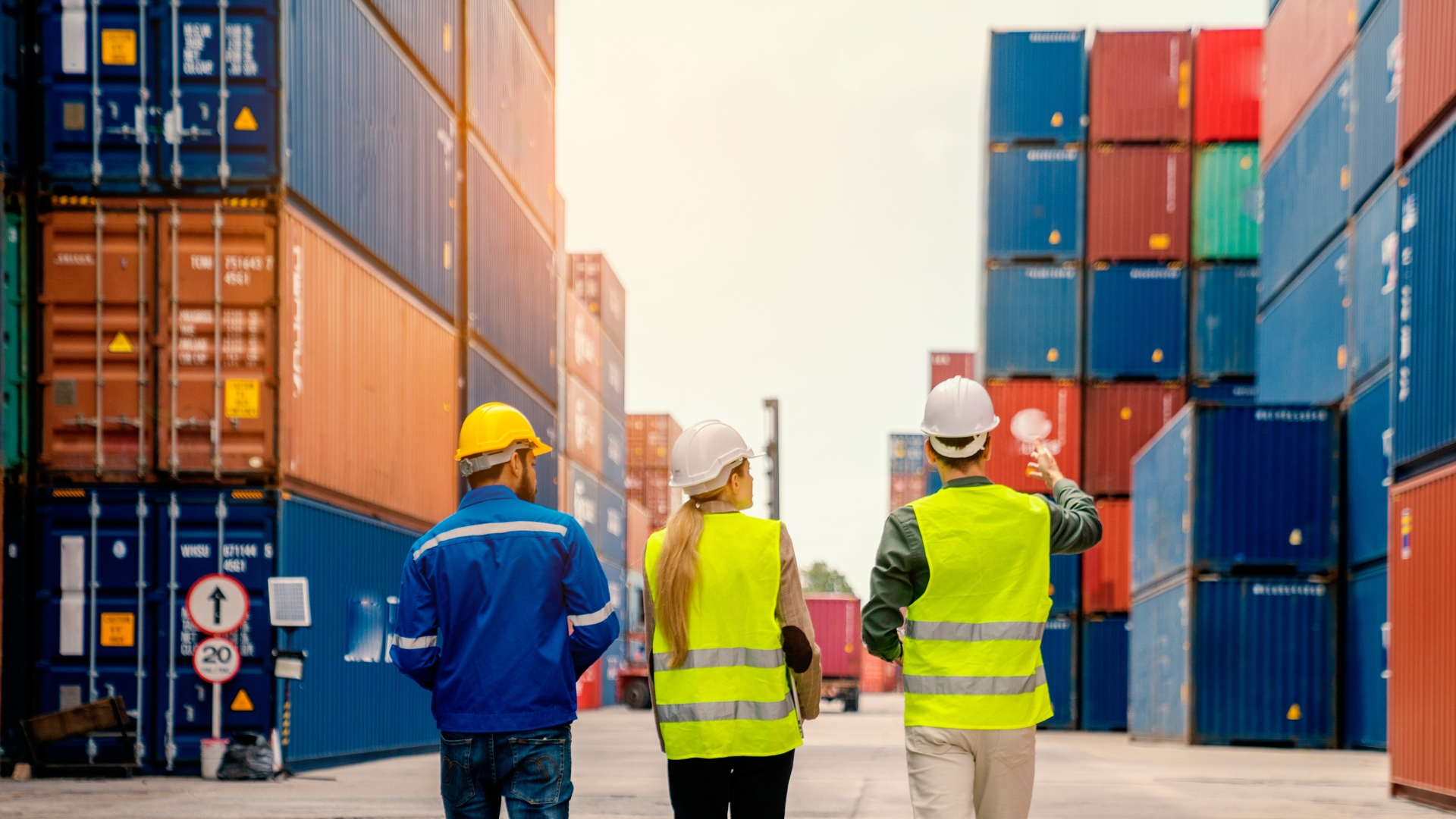Three workers wearing safety helmets and reflective vests inspect stacked shipping containers at a busy port, symbolizing global trade and import logistics.