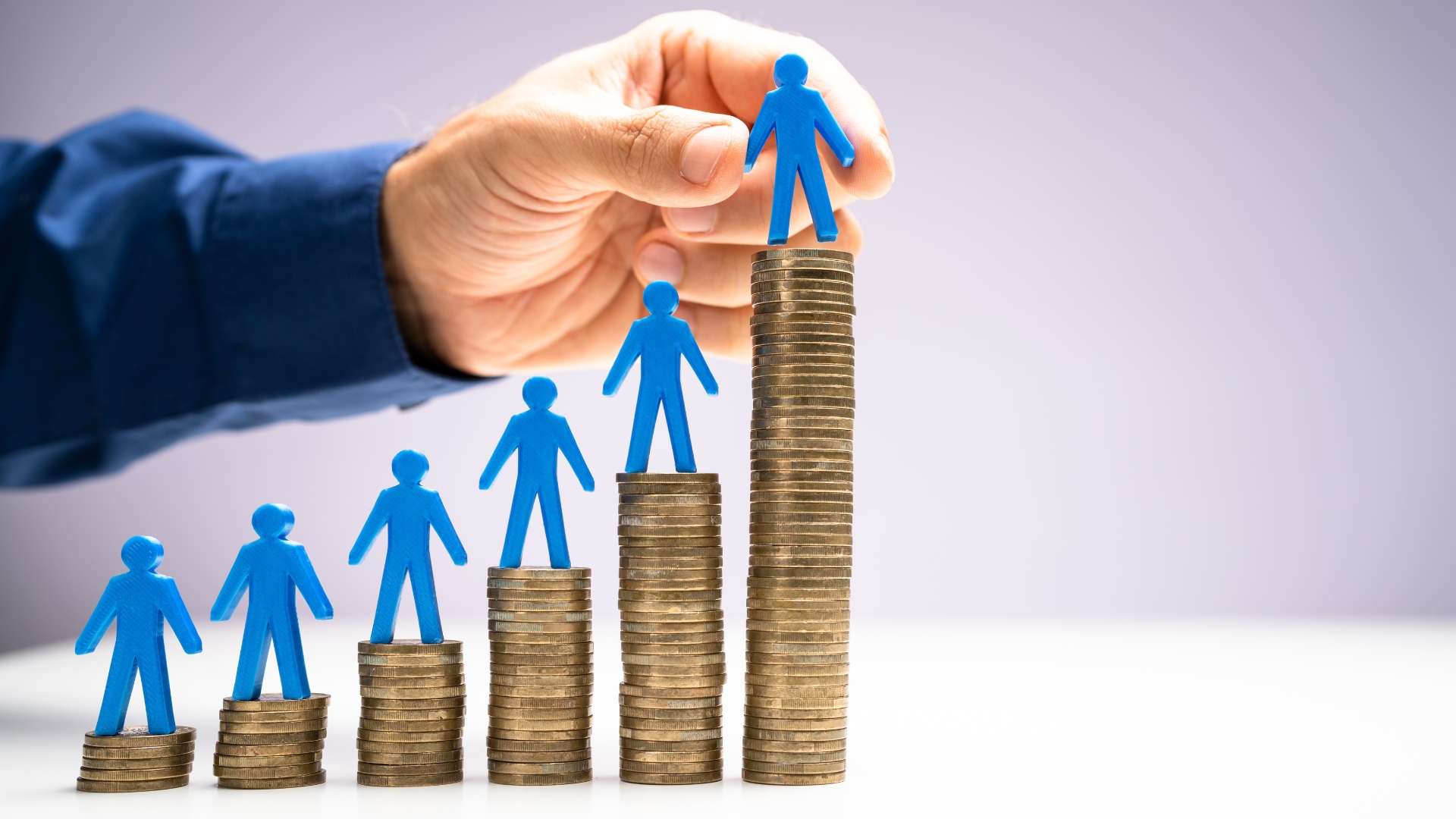 Blue human figures standing on ascending stacks of coins representing employee ownership and growing financial benefits, with a hand placing the top figure on the highest stack.