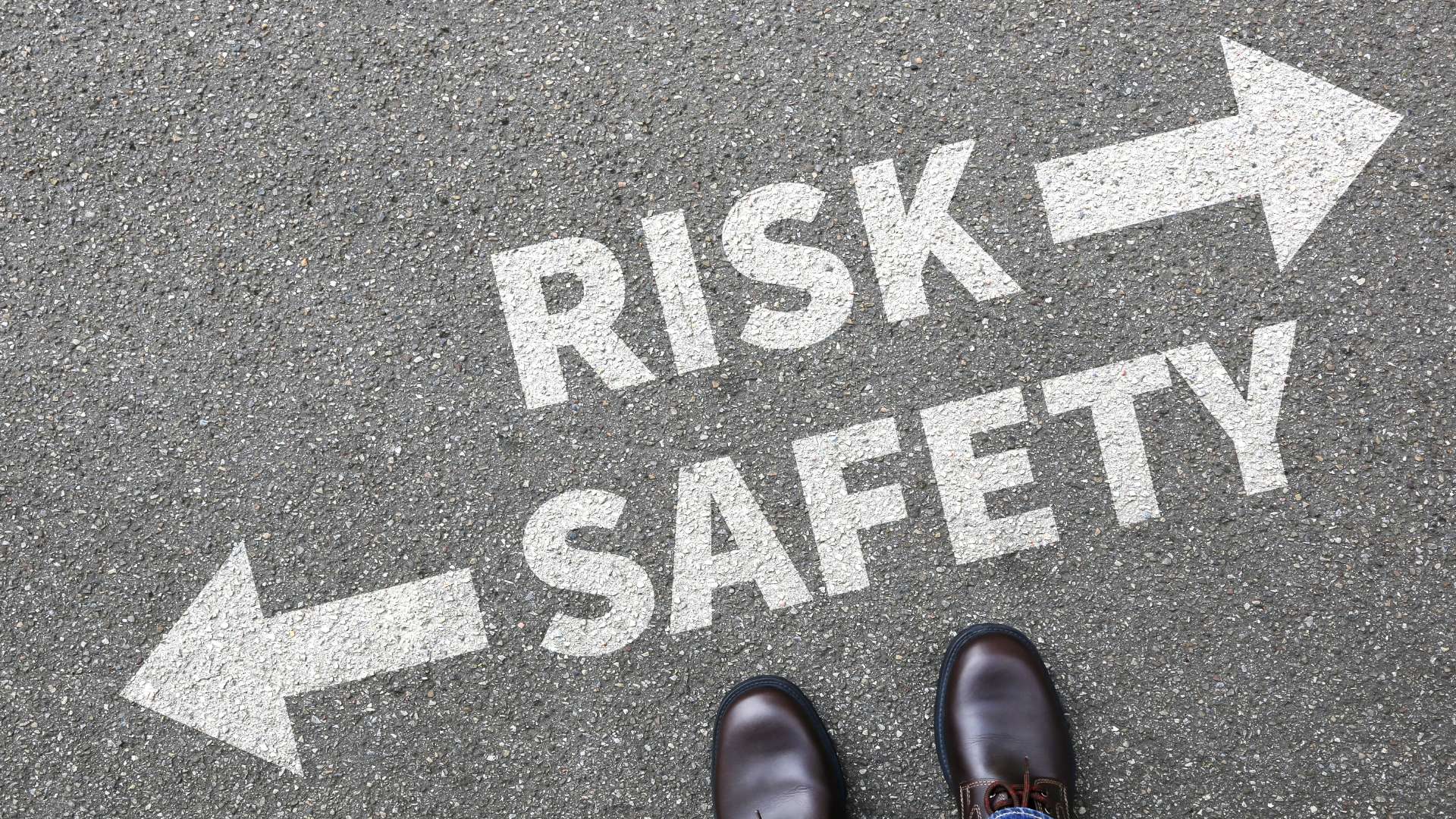 A high-angle view of a person's feet in leather shoes standing on asphalt between two painted white arrows pointing in opposite directions, one labeled "RISK" and the other labeled "SAFETY."
