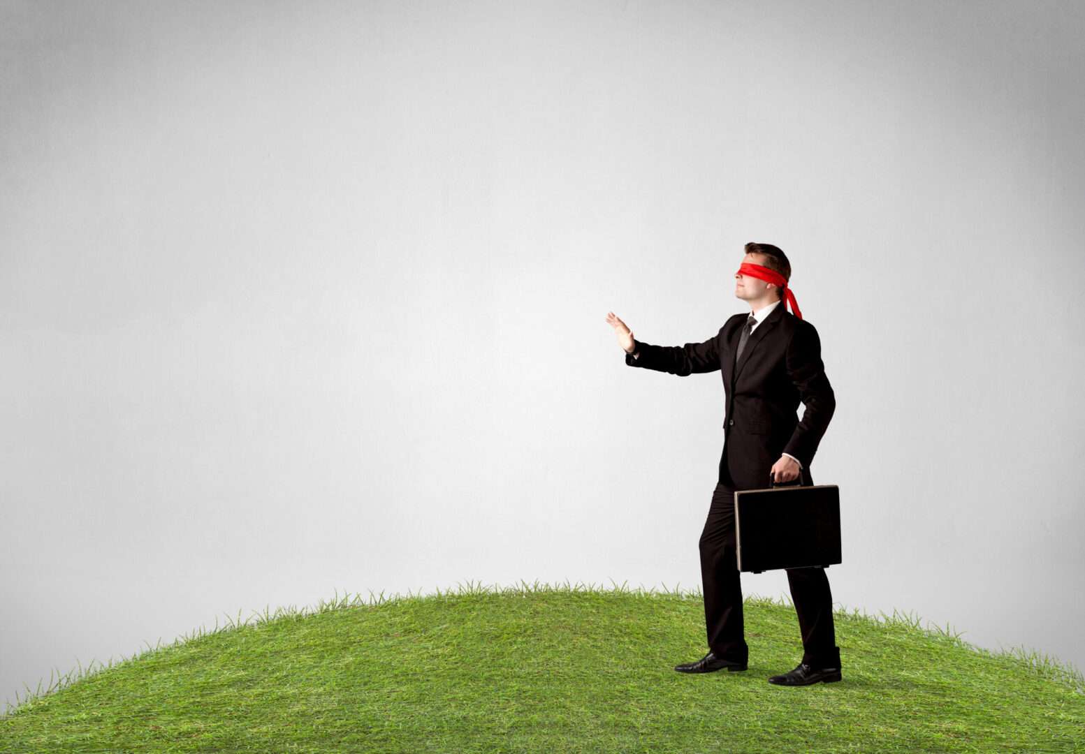 A blindfolded businessman in a suit carrying a briefcase, walking cautiously on a patch of grass, representing an entrepreneur navigating trademark laws without legal guidance.