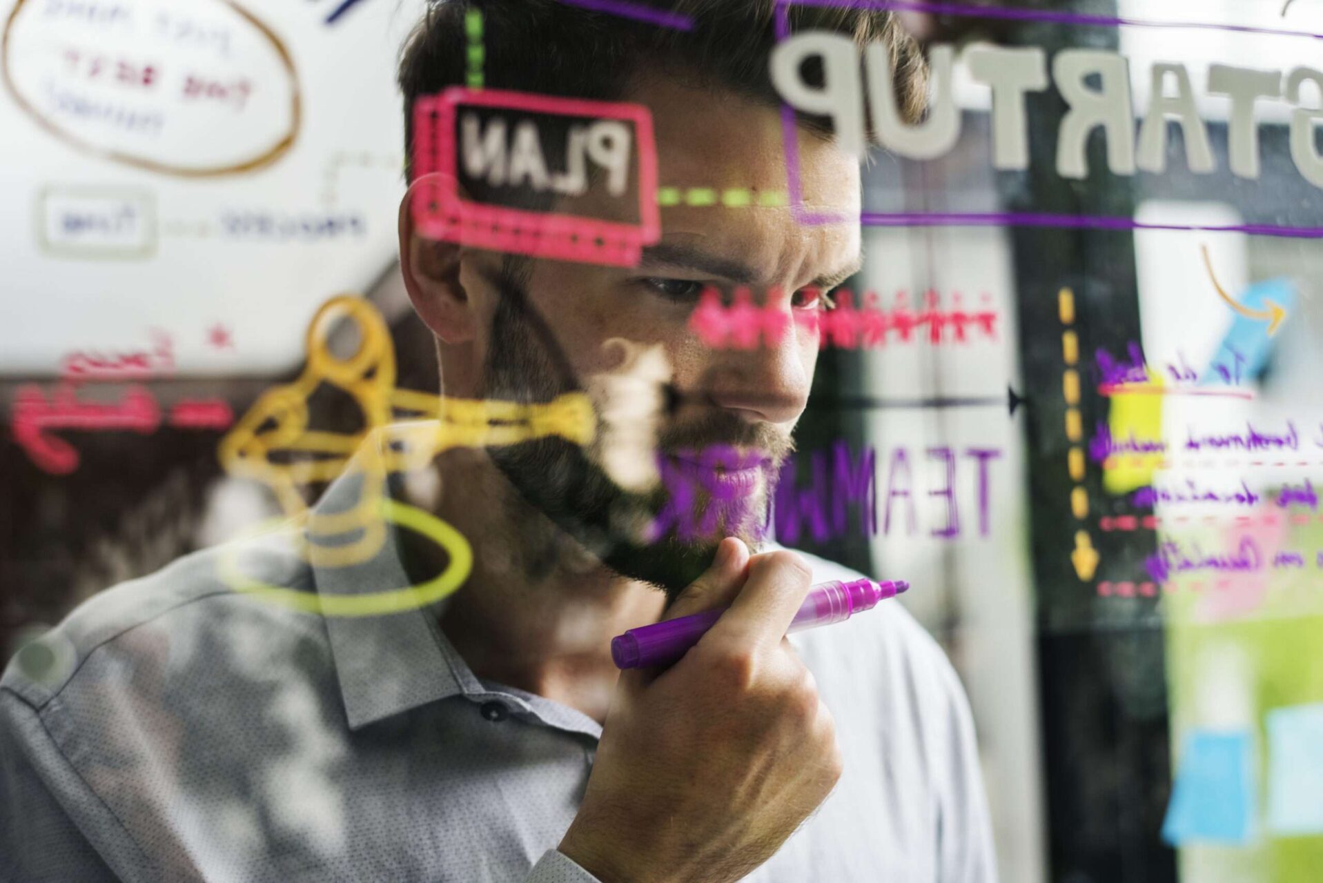A business professional brainstorming startup branding ideas on a glass board, with colorful markers showing words like "Plan," "Startup," and "Teamwork," representing the strategic process of choosing between a logo and wordmark for trademark protection.