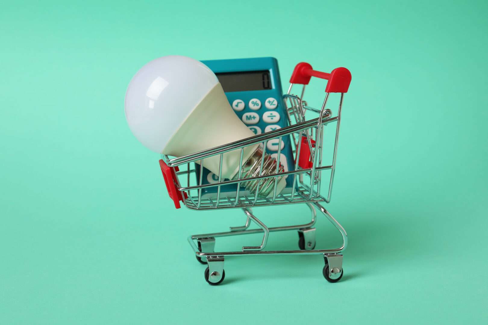 A miniature shopping cart containing an LED light bulb and a calculator on a mint background, representing the smart financial and intellectual property decisions e-commerce business owners must make to protect their brand assets.