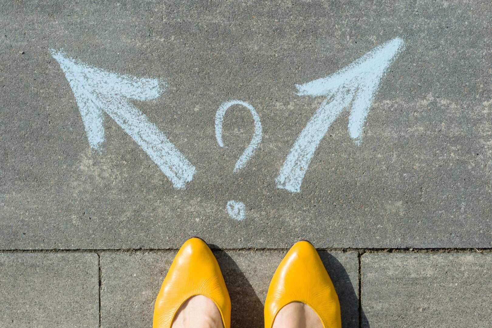 A person in yellow shoes standing on pavement facing two chalk arrows pointing in opposite directions with a question mark in the middle, symbolizing the confusing choice between a business name registration and a federal trademark.