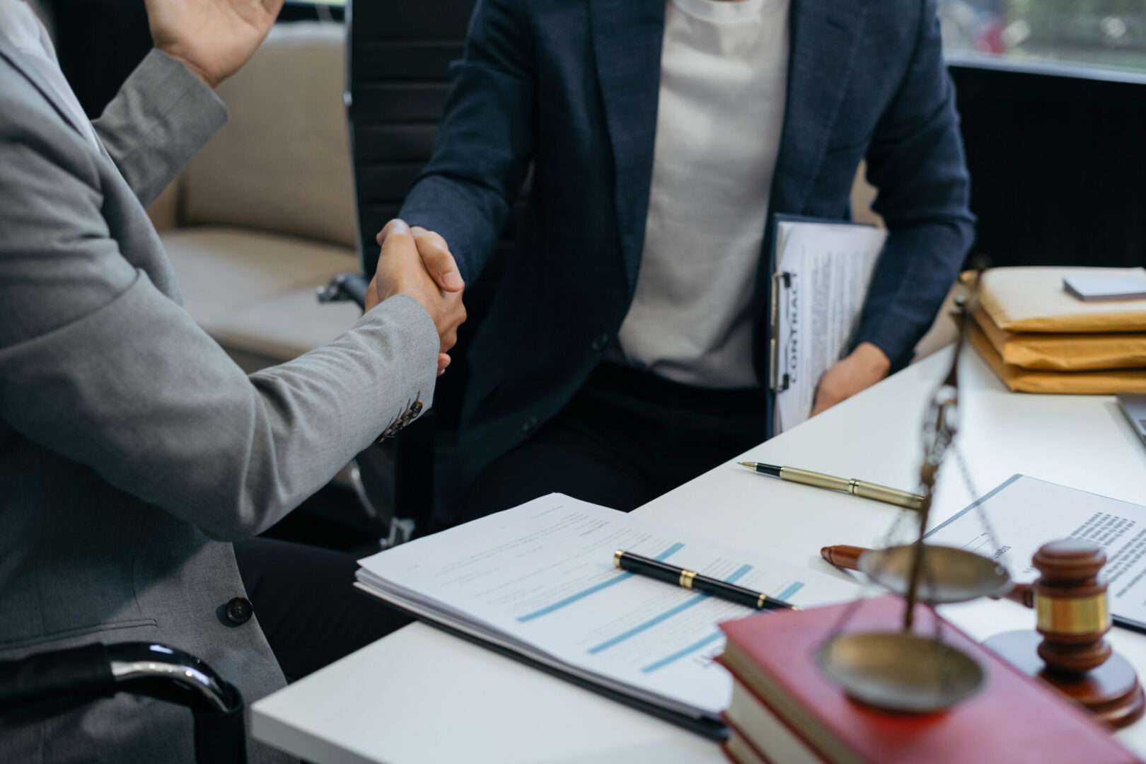 Two business professionals shaking hands in a legal office setting with a contract clipboard, scales of justice, gavel, legal documents, and gold pens on the desk, representing the trusted partnership between a small business owner and a Virtual General Counsel at Carbon Law Group.