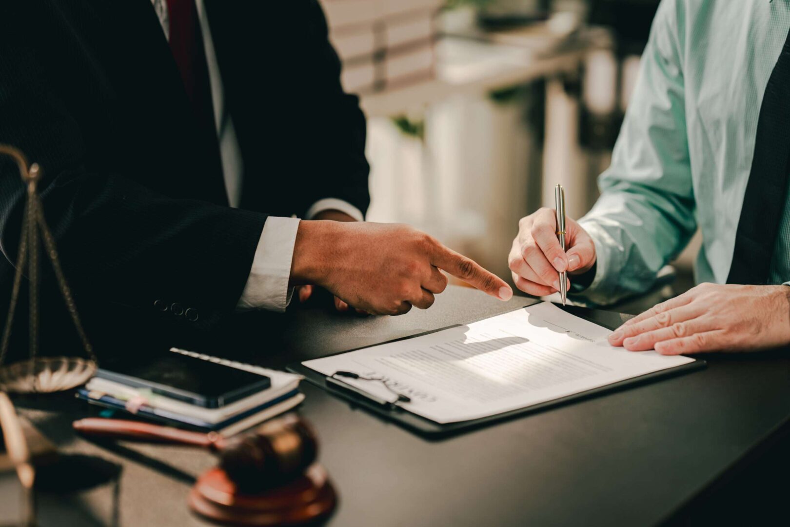 An attorney pointing to a specific clause in a legal document while a client signs, with a gavel and scales of justice visible on the desk, representing the IP licensing agreement review and trademark protection services Carbon Law Group provides for small business owners in Los Angeles.