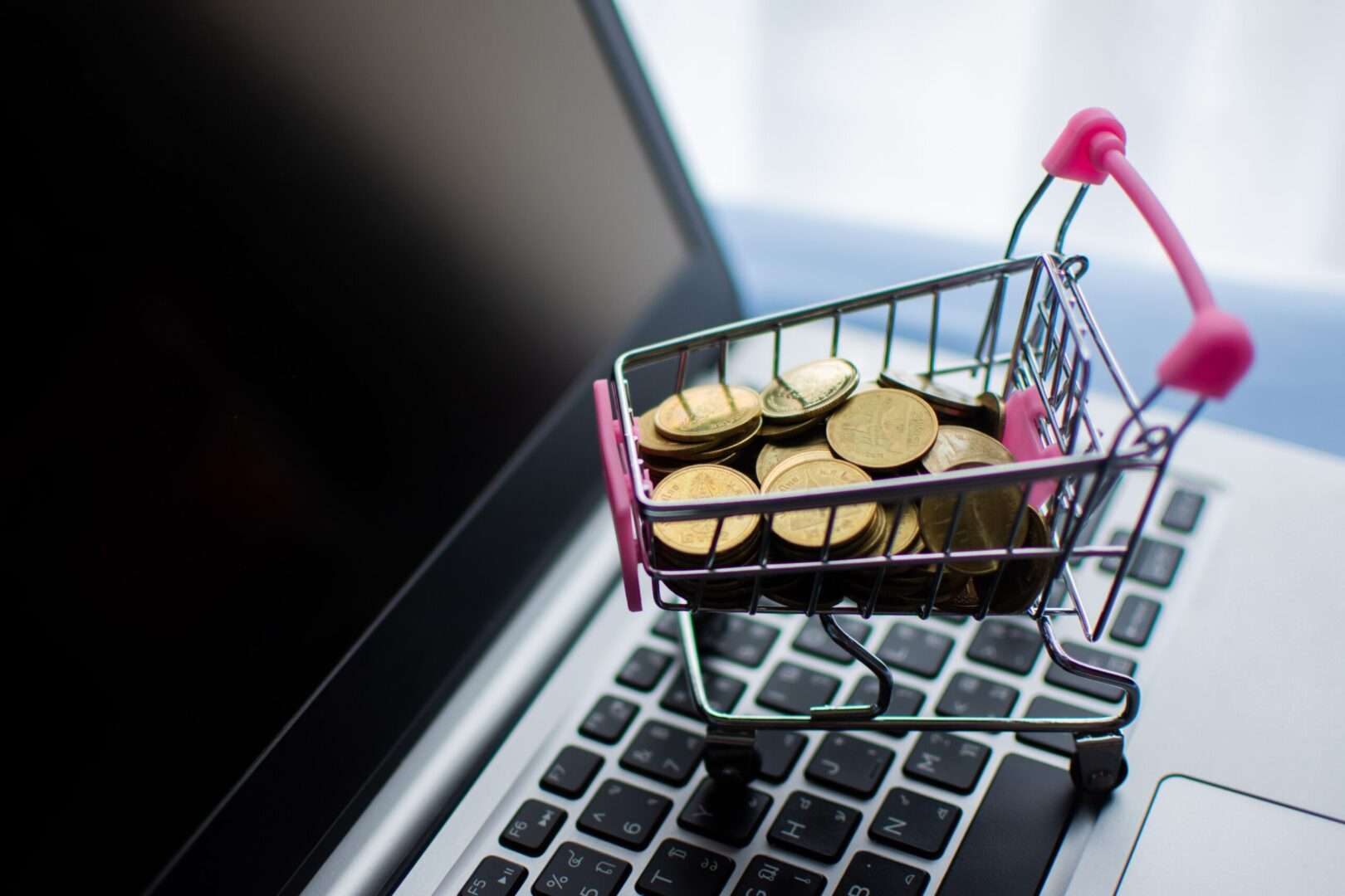 A miniature shopping cart filled with gold coins sitting on a laptop keyboard, representing the financial value and revenue protection that ongoing legal support provides for direct-to-consumer brands in Los Angeles.