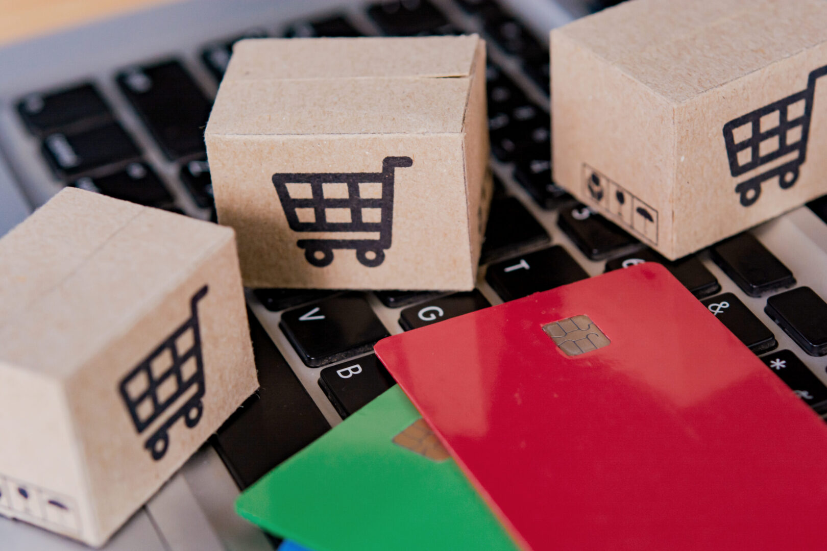 Small cardboard boxes printed with shopping cart icons alongside red and green credit cards on a laptop keyboard, representing the legal risks and transactional complexity that e-commerce brands in Los Angeles must navigate with Fractional General Counsel support.