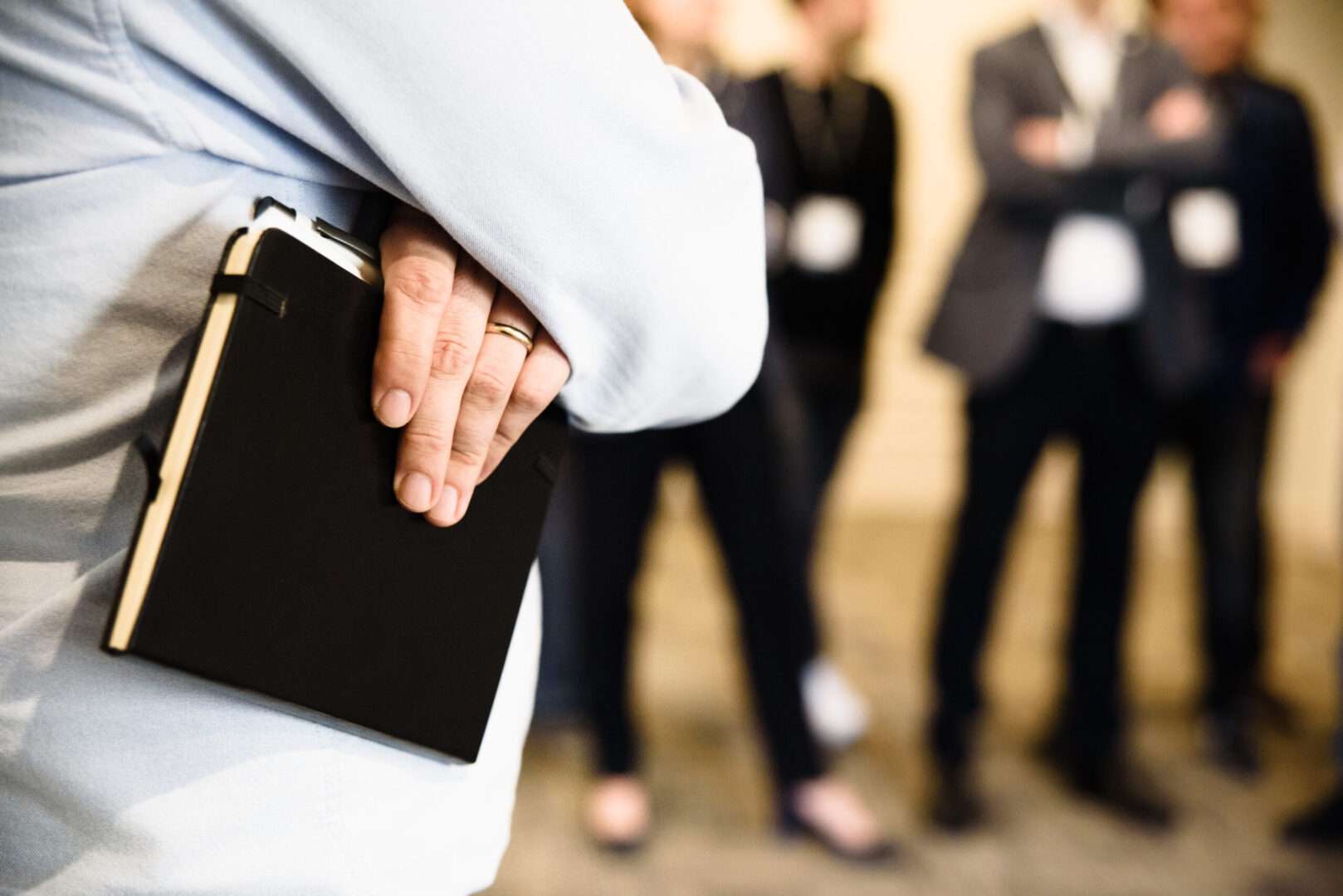 A professional holding a black notebook at a business conference with colleagues in the background, representing the importance of documentation, clear workplace policies, and written agreements that protect small business owners from costly employment disputes.