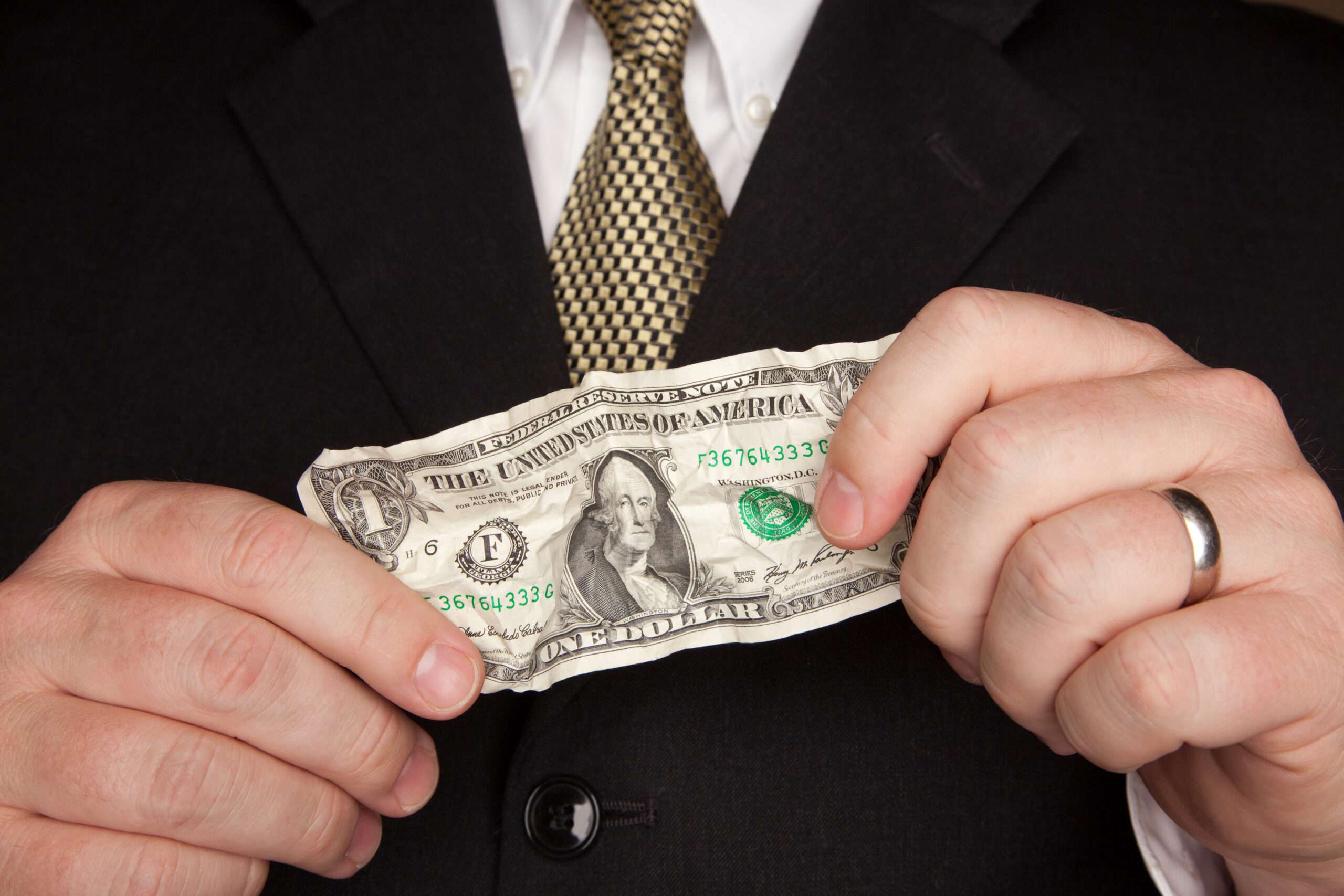 A business professional in a dark suit and gold tie smoothing out a crumpled one dollar bill, representing the cost decisions businesses weigh when choosing between a legal subscription and hiring in-house counsel.