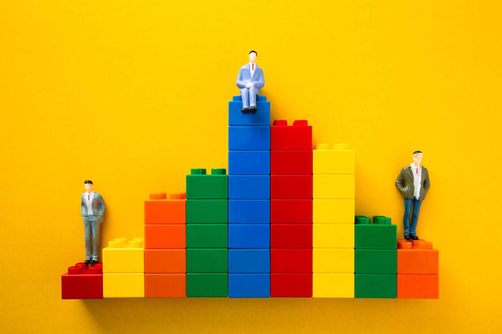 Miniature businessman figurines stand and sit on a colorful staircase of building blocks against a yellow background, representing the steady, structured growth of a DTC brand scaling in California.