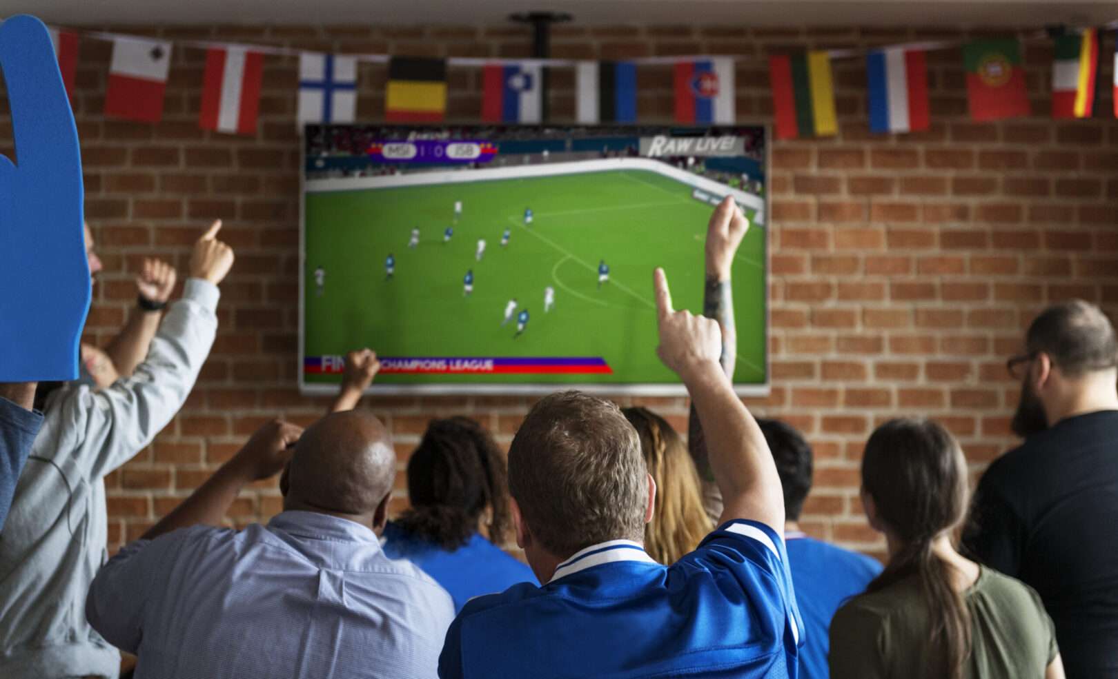 A crowd of cheering fans points at a big-screen TV showing a live soccer match in a bar decorated with international flags, representing the 2026 FIFA World Cup viewing crowds that LA small businesses can legally capitalize on.