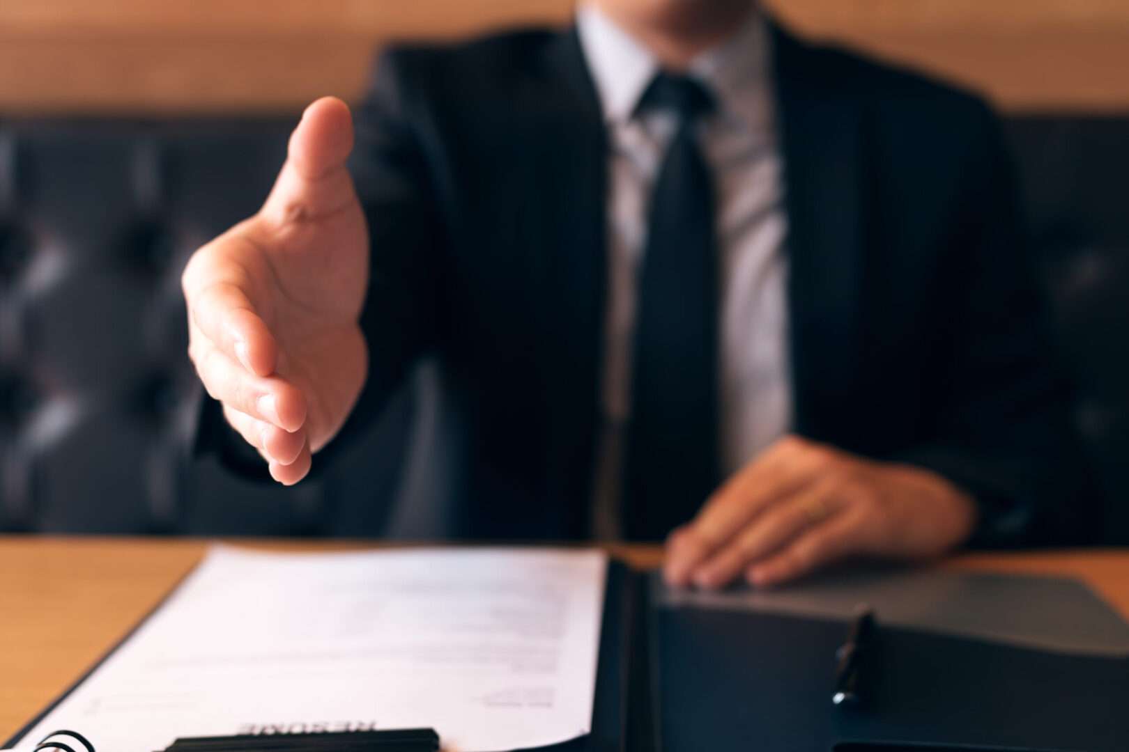 A business professional in a dark suit extends a hand for a handshake across a desk with documents and a folder, representing the proactive legal partnership $5M revenue companies in LA need to protect their growth.
