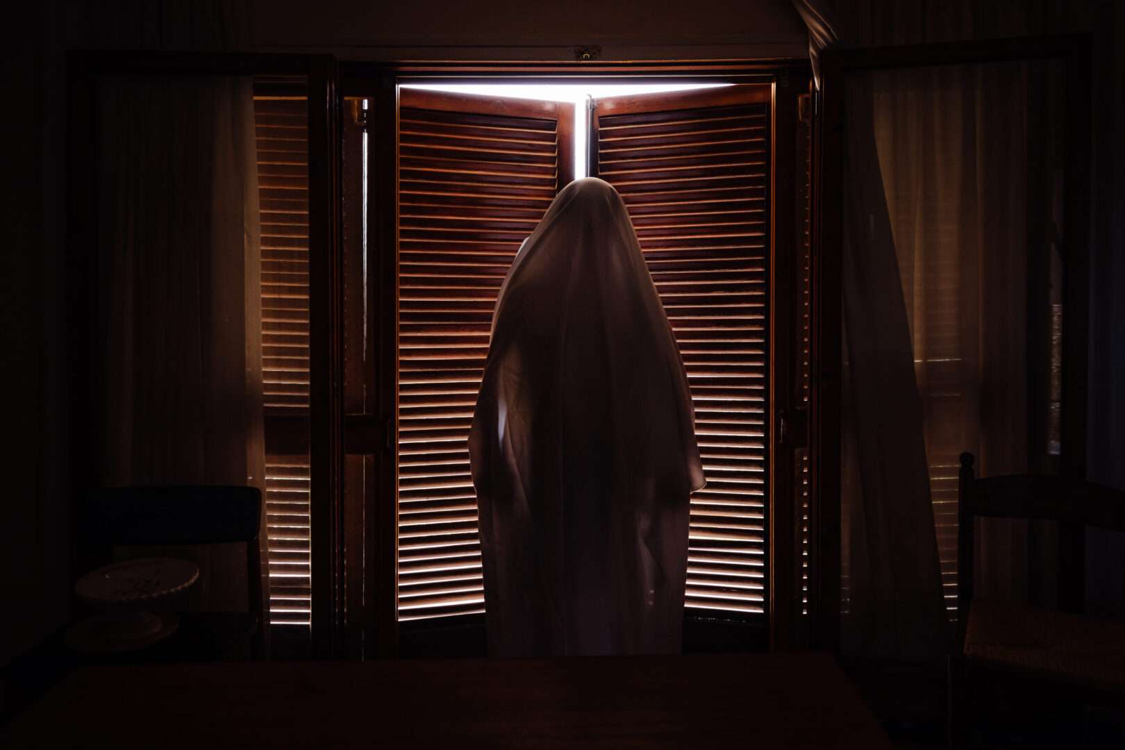 A figure draped in a white sheet like a classic ghost stands silhouetted against wooden window shutters in a dimly lit room, evoking the supernatural horror film "Obsession" and the IP lessons it offers creators and small businesses.