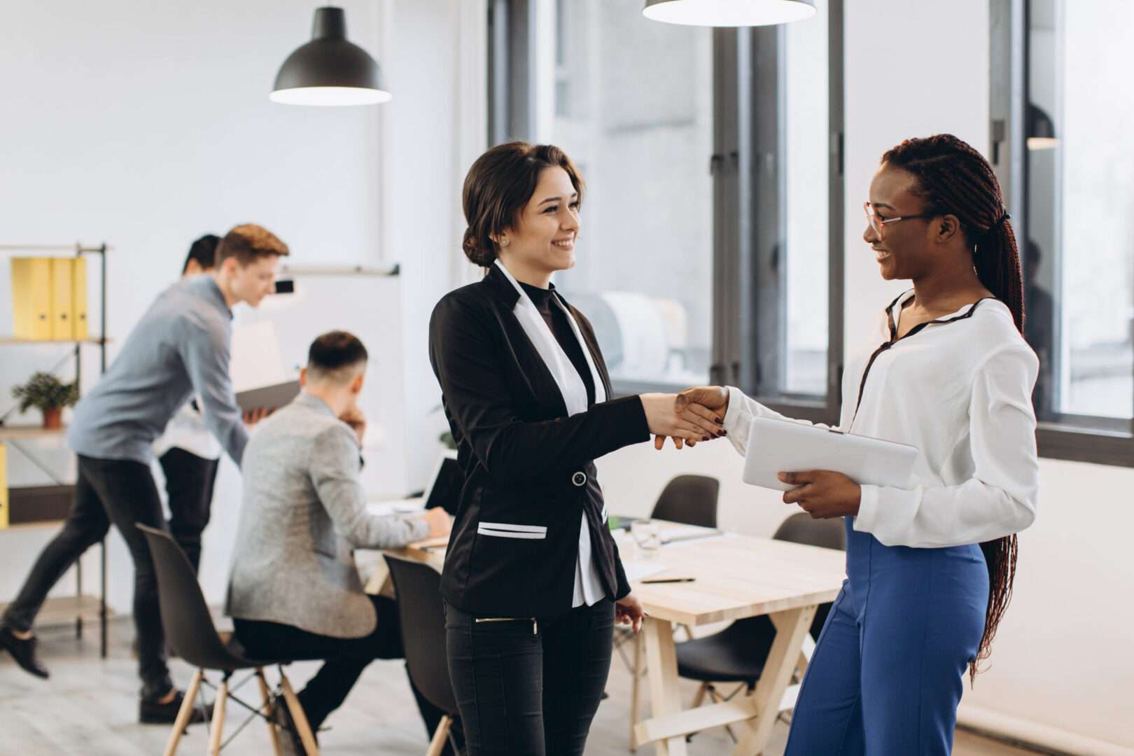 Two businesswomen smile and shake hands in a modern office while colleagues work in the background, representing the trust and professionalism that come from keeping a California LLC compliant.
