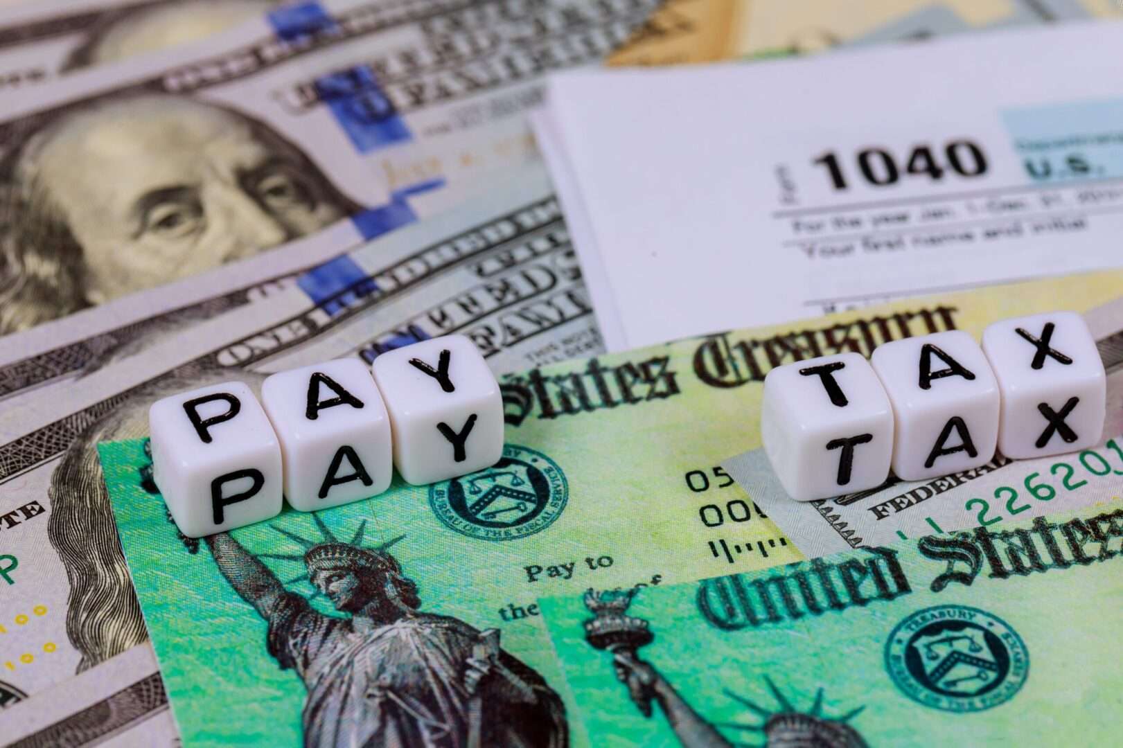 White letter dice spelling "PAY TAX" sit on top of US Treasury refund checks, hundred dollar bills, and a Form 1040, representing the tax differences between a California LLC and an S-Corp for online businesses.