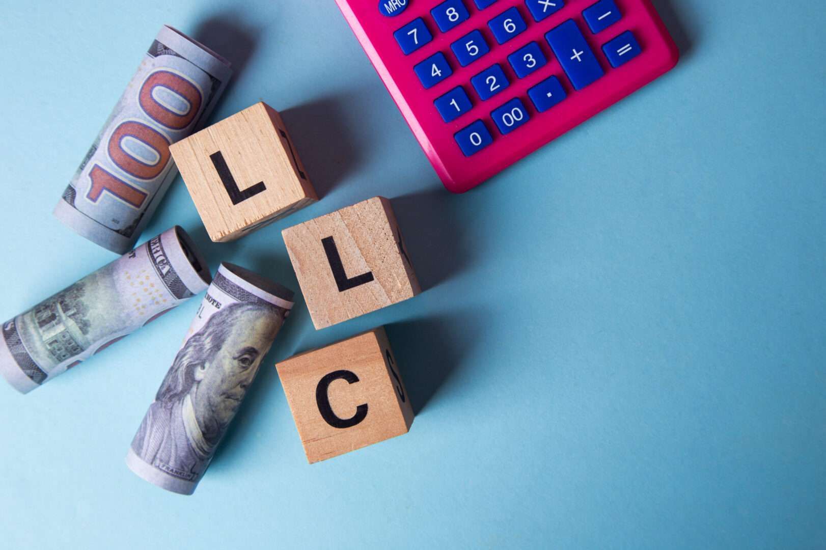 Wooden letter blocks spelling "LLC" sit beside rolled one hundred dollar bills and a pink calculator on a blue background, representing the process and costs of forming an LLC for a California e-commerce business.