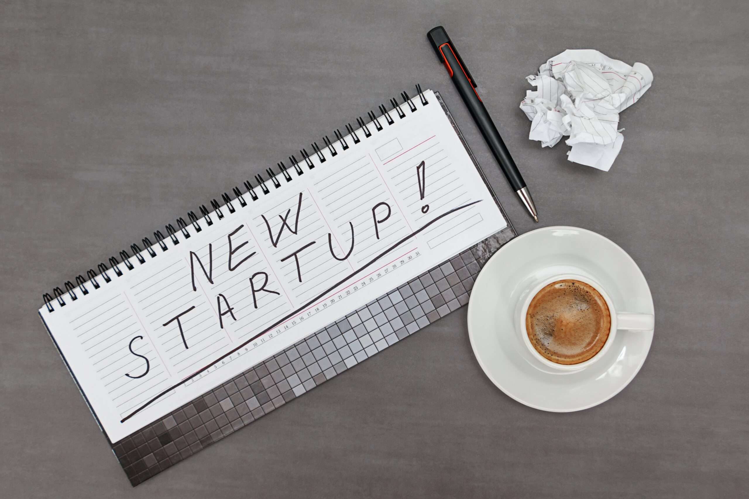 A spiral notepad with "New Startup!" handwritten across it sits beside a pen, a crumpled sheet of paper, and a cup of espresso on a gray desk, representing the early planning stage of forming a new business in LA.