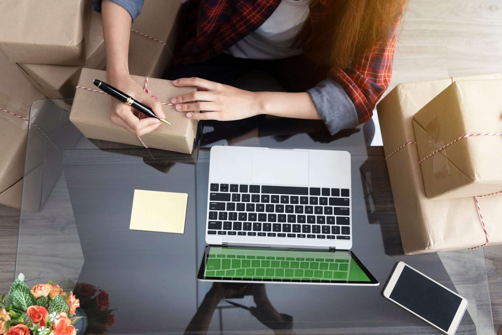 An online business owner labels shipping boxes at a glass desk with a laptop, sticky note, and phone, representing a startup founder deciding between a Delaware C-Corp and a California LLC.
