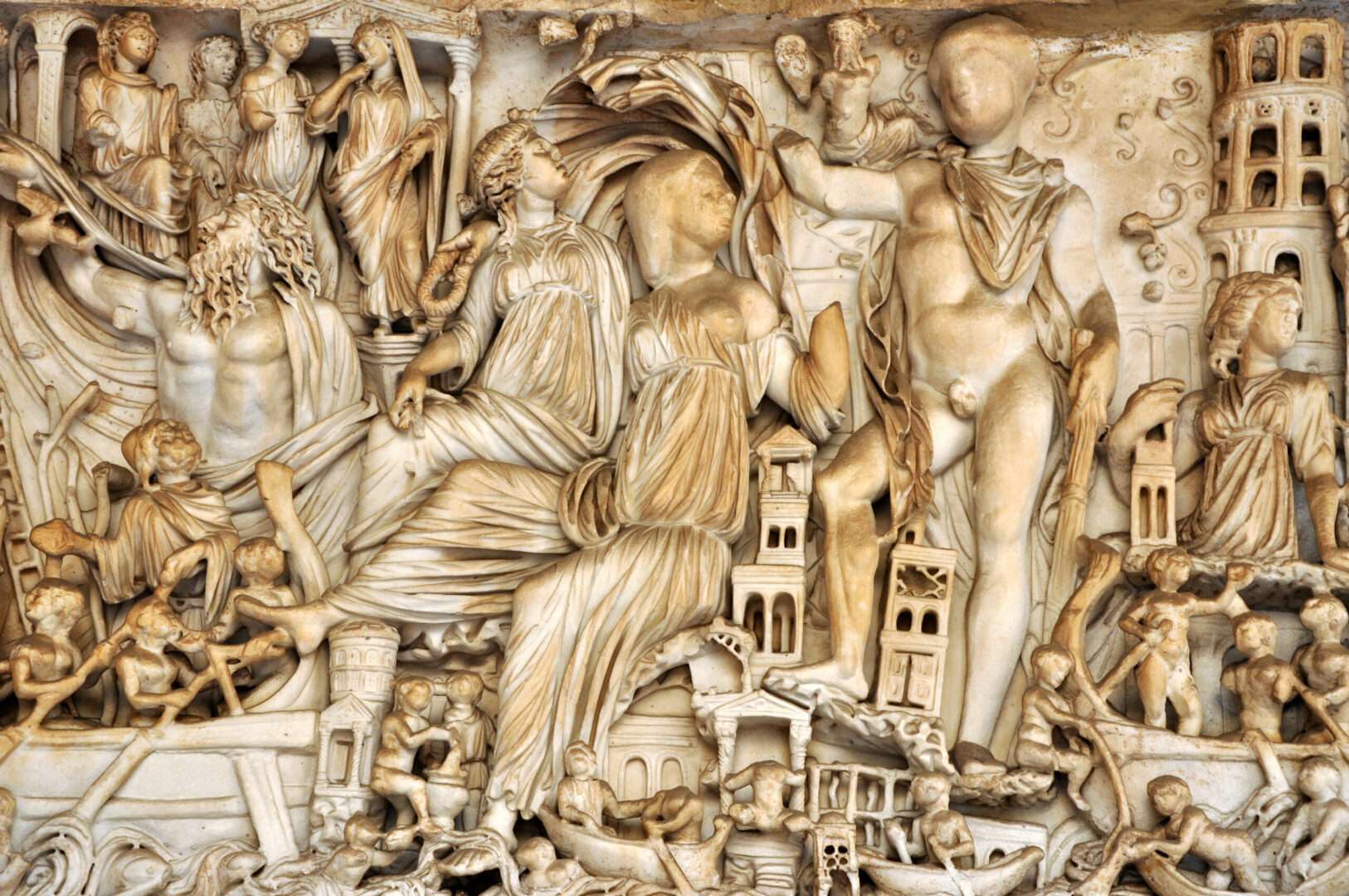 An ornate classical stone relief carved with mythological figures, boats, and towers, evoking the epic source material behind Christopher Nolan's film "The Odyssey."