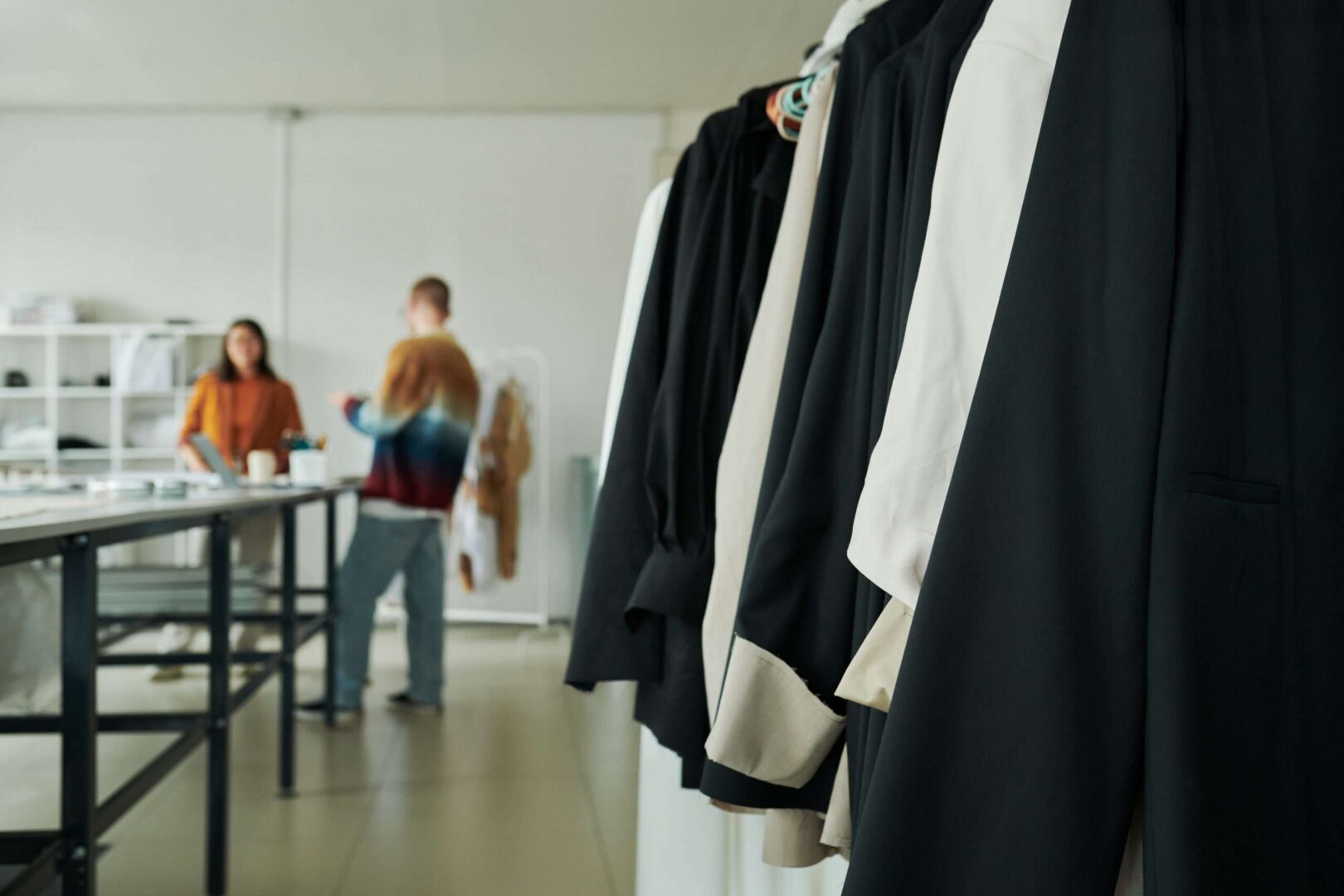 A rack of elegant black and beige jackets hanging in a fashion design studio while two creative entrepreneurs work together in the background.