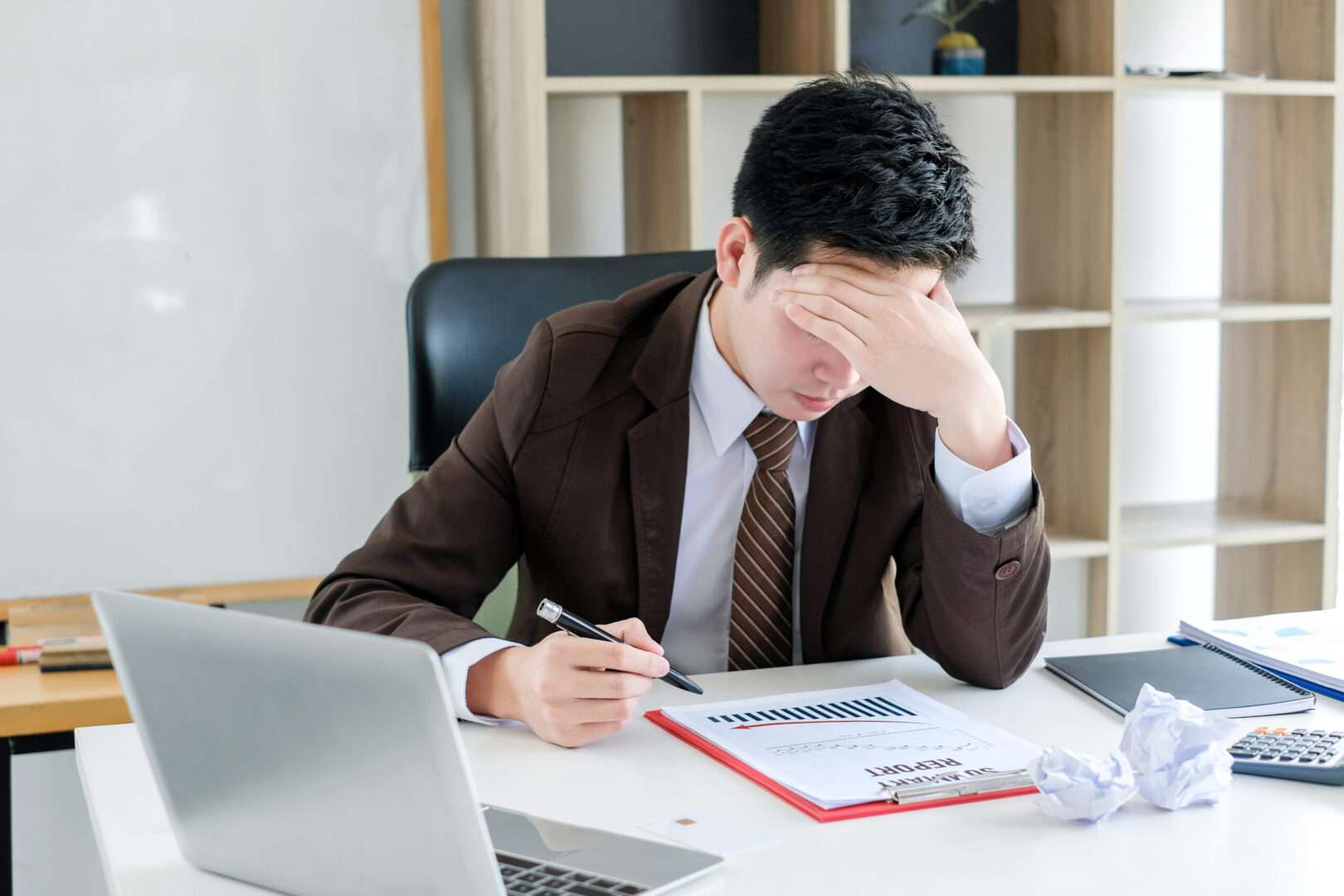 A business owner rests his head in his hand at a desk with a laptop, a summary report, and crumpled paper, representing the stress and disputes that arise without a solid operating agreement.