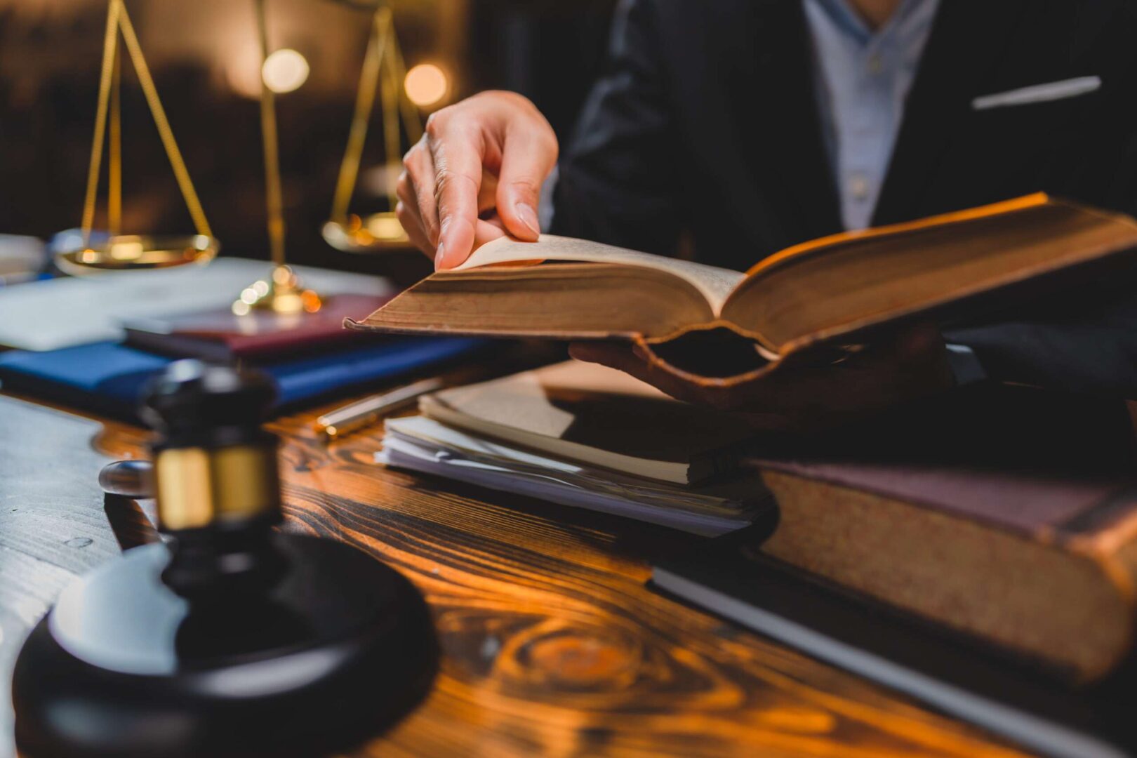An attorney turning the pages of an open law book at a wooden desk with a gavel and golden scales of justice.