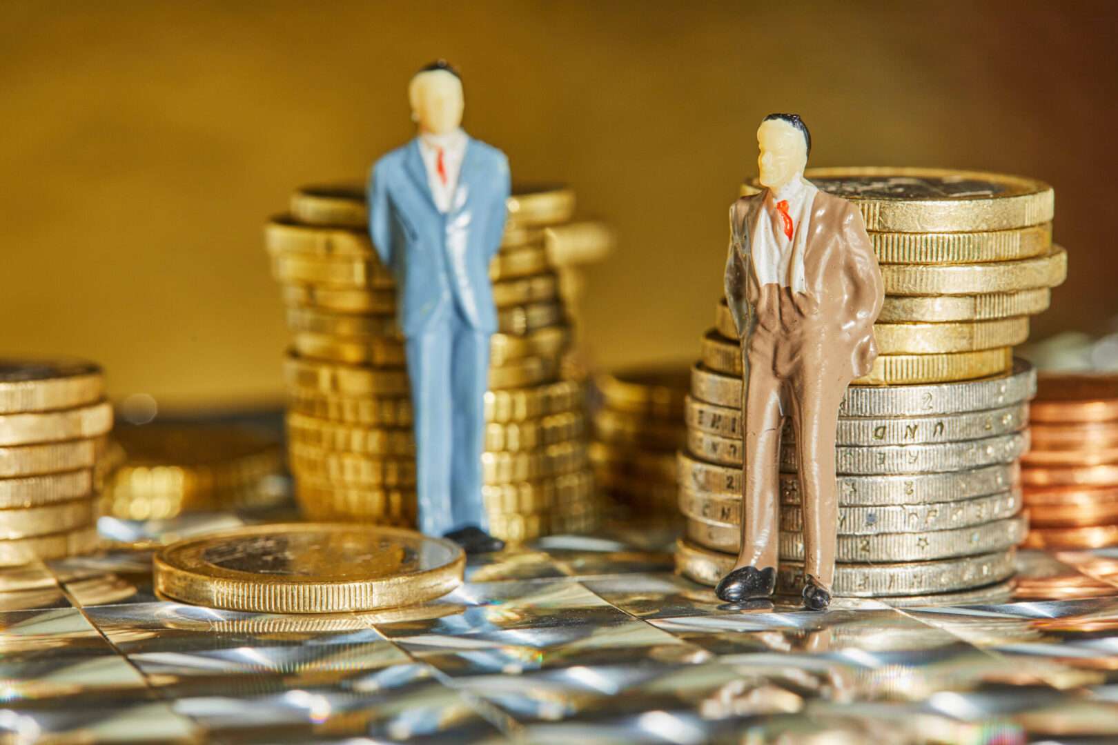 Two miniature businessman figures stand beside stacks of coins of different heights on a reflective surface, representing majority and minority owners and the equity divide between business partners.