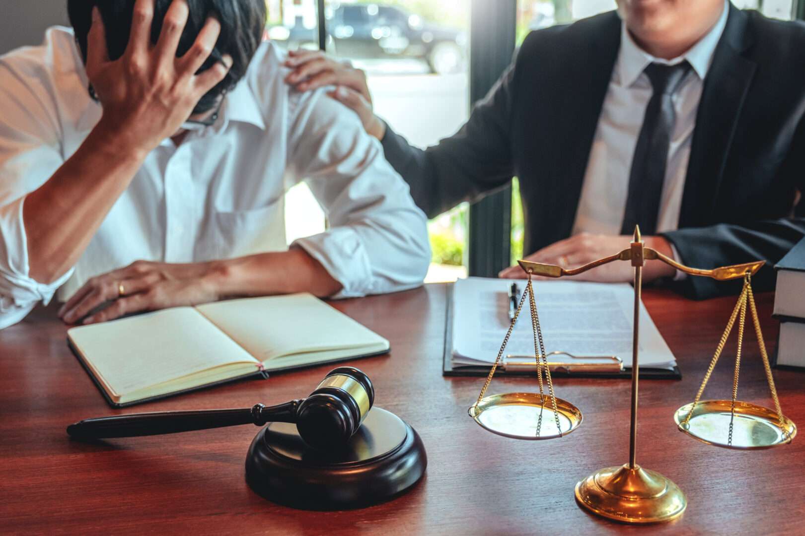 A stressed business founder holding his head in his hands while an attorney in a suit places a supportive hand on his shoulder at a desk with legal scales and a gavel.