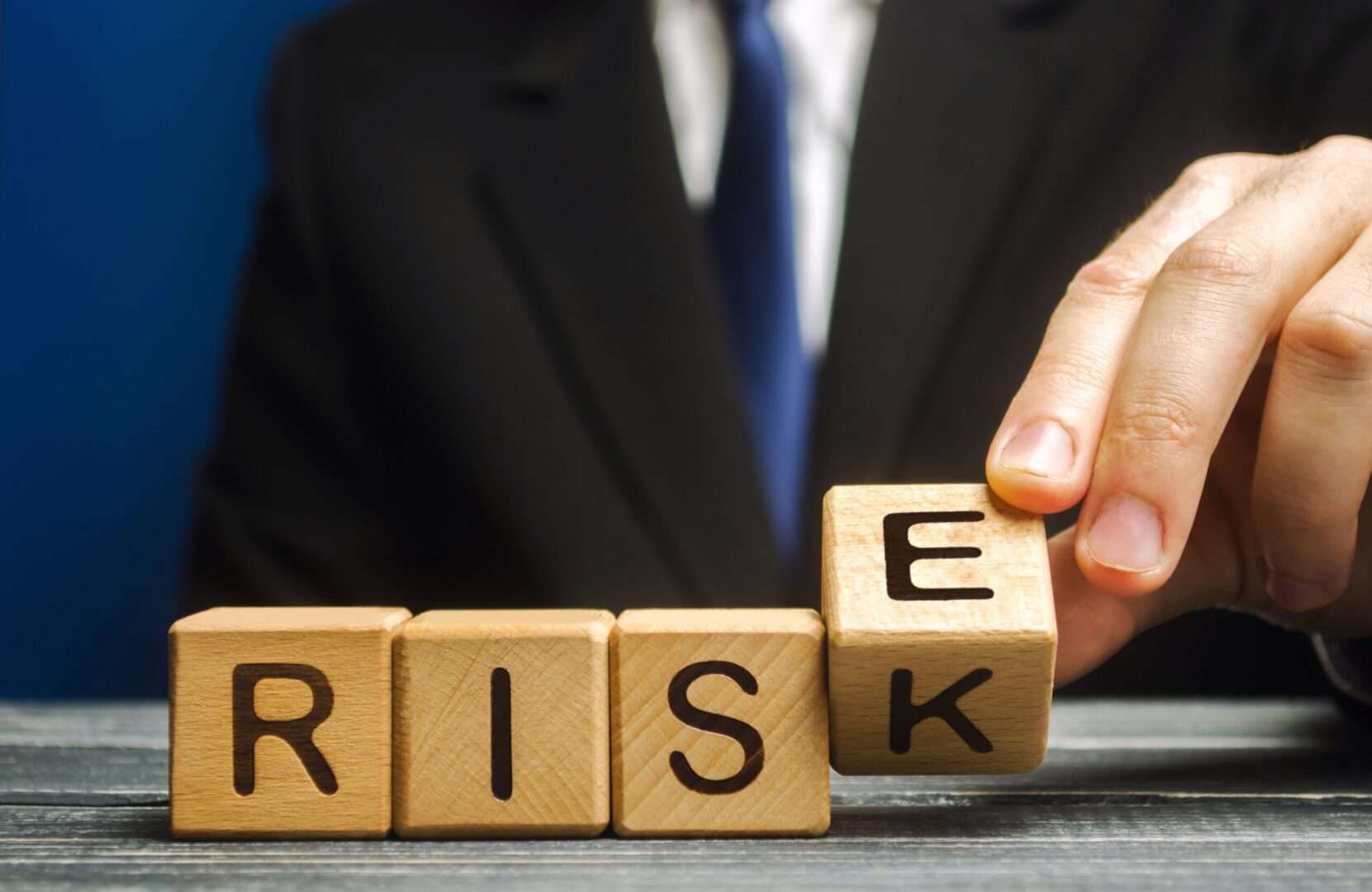 A business professional turning a wooden letter block to change the word "RISK" to "RISE" on a dark wooden office desk.
