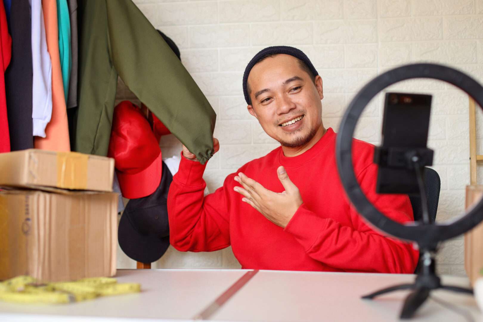 A male fashion e-commerce entrepreneur filming a live product showcase on his smartphone in a home studio next to clothing racks and shipping boxes.