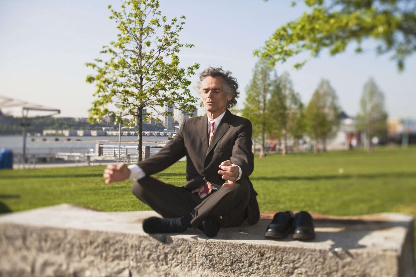 Business professional in a suit meditating cross-legged on a stone bench in a waterfront park, illustrating mindful approaches to founder stress