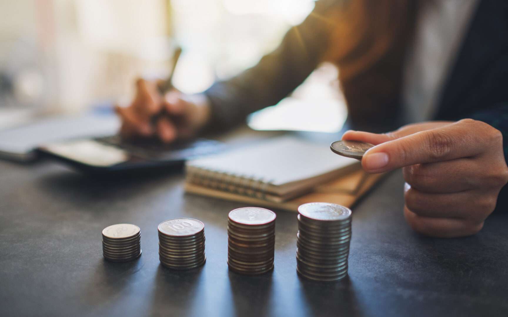 A business owner stacking coins into neat piles on an office desk next to a notebook, pen, and calculator to manage business finances.