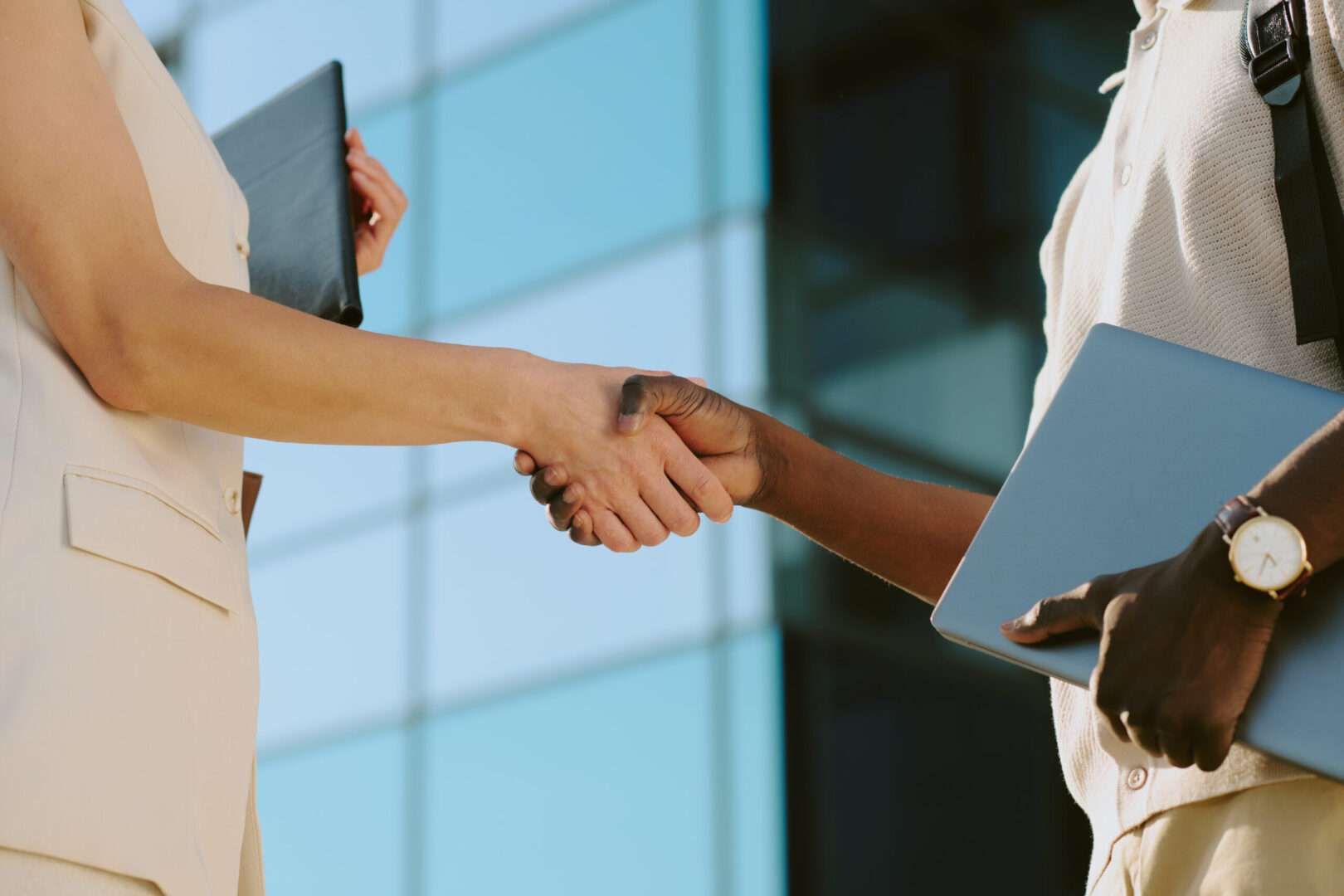 Two business professionals shake hands outside a modern glass office building while holding a laptop and portfolio, representing a vendor partnership backed by a solid agreement.