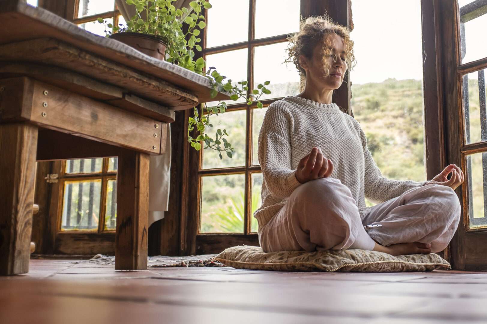 Woman sitting cross-legged on a floor cushion by a large window, practicing her daily meditation habit in a sunlit room