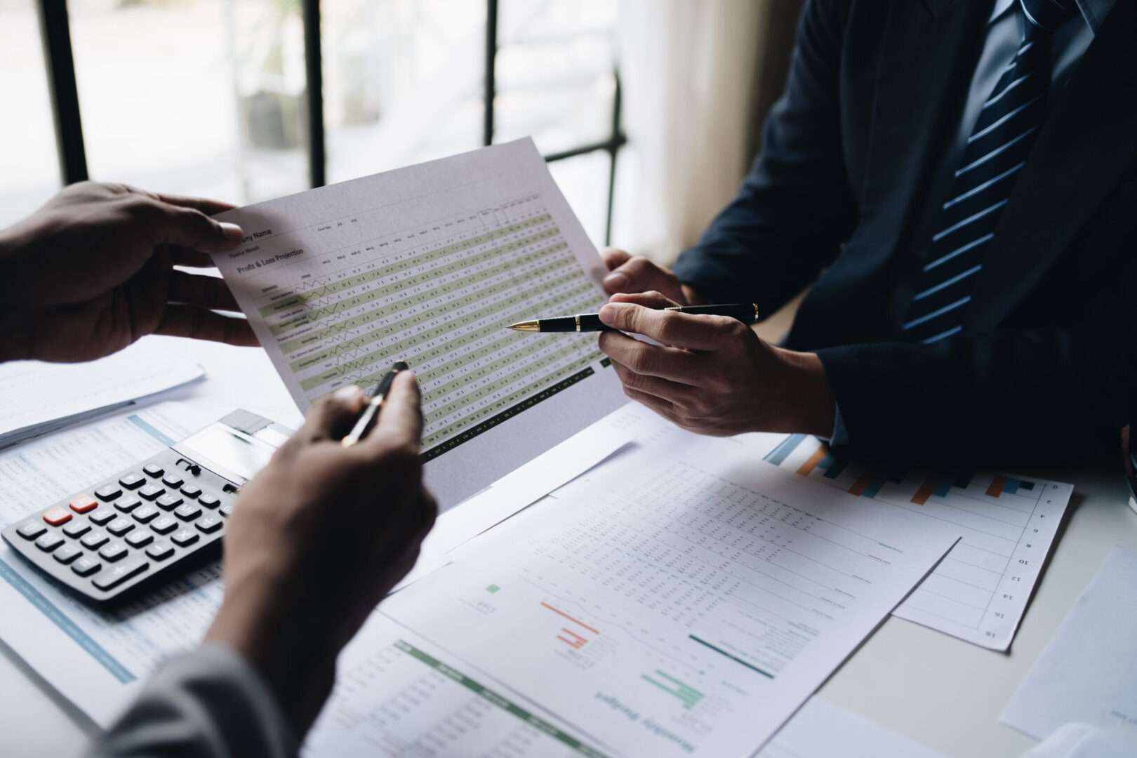 Two colleagues reviewing a twelve-month profit and loss projection with a calculator and financial reports on the desk