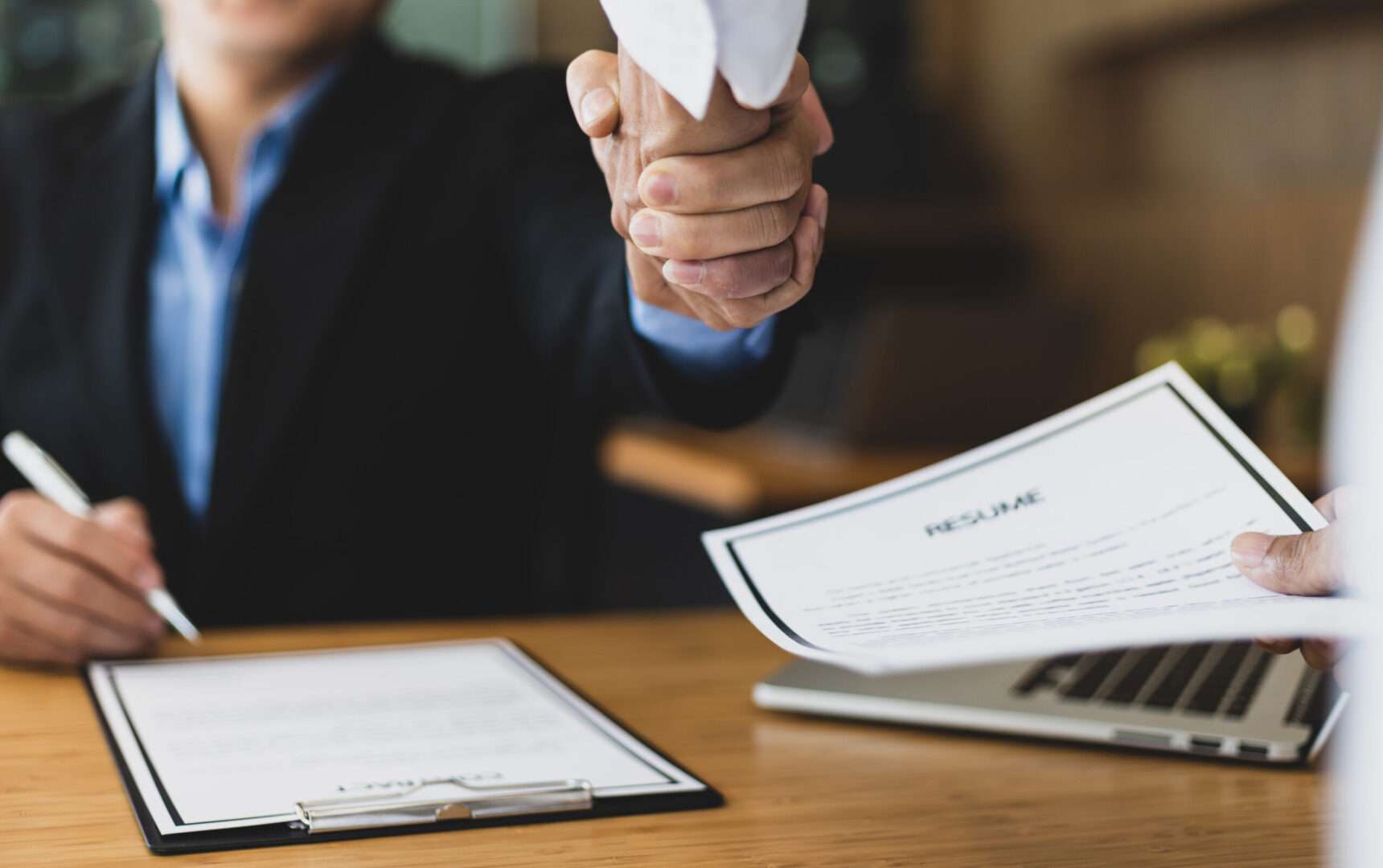 Two people shaking hands across a desk over a contract clipboard, laptop, and printed documents