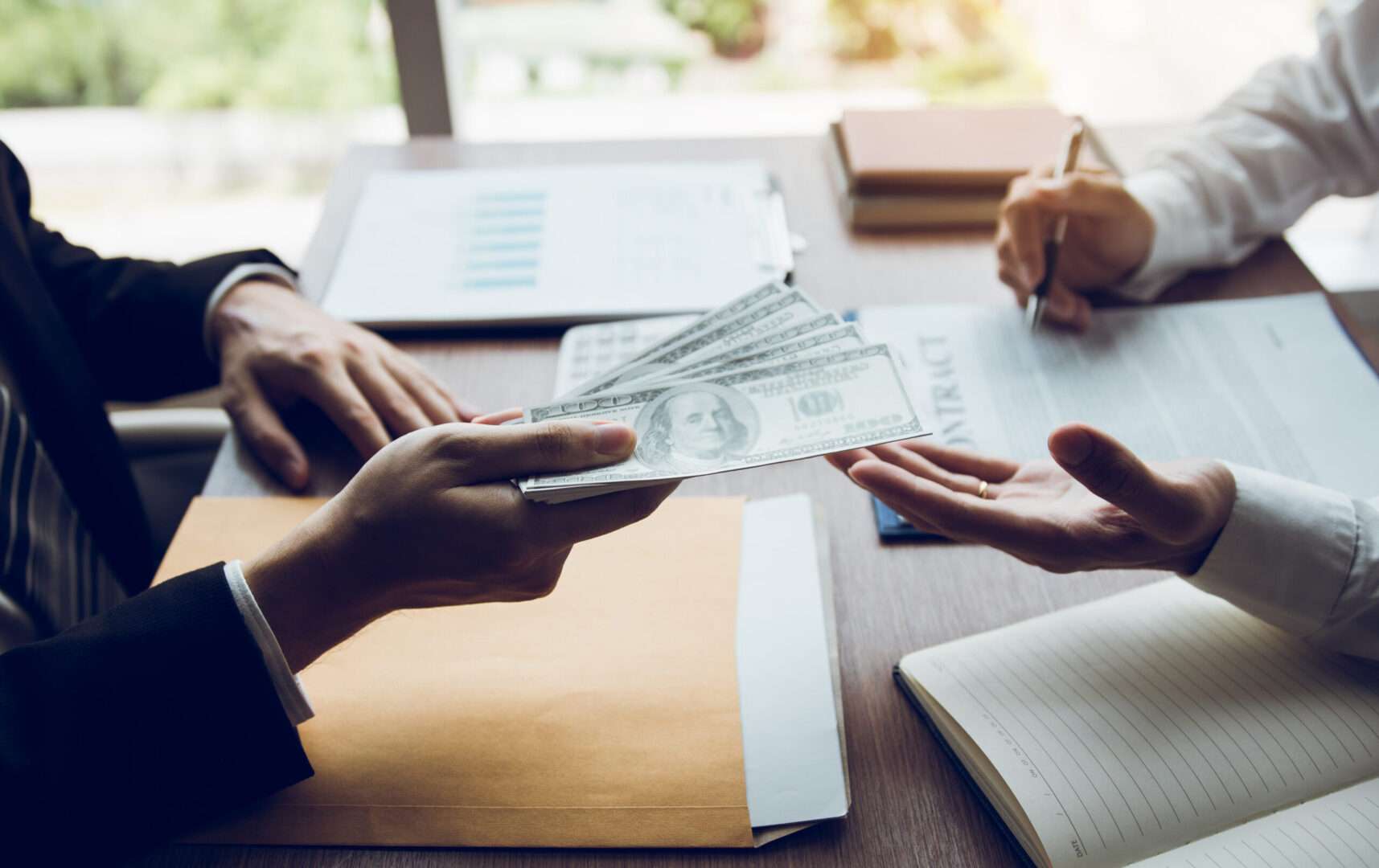 One person hands a fan of hundred dollar bills across a desk while another signs a contract, representing payment in a business sale