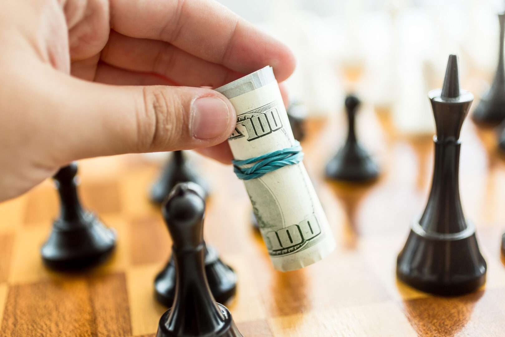 Hand placing a roll of hundred dollar bills among chess pieces, representing a seller reinvesting proceeds as rollover equity