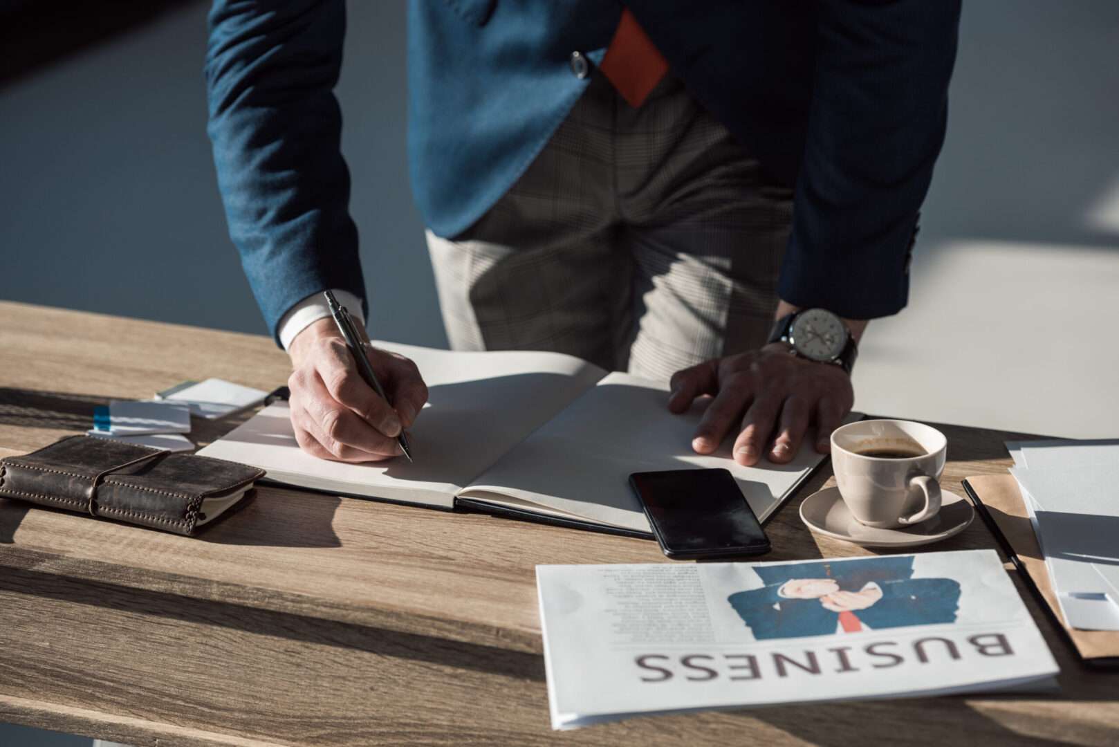 Founder writing in a notebook at a desk with a business newspaper, phone, and coffee while planning a funding round