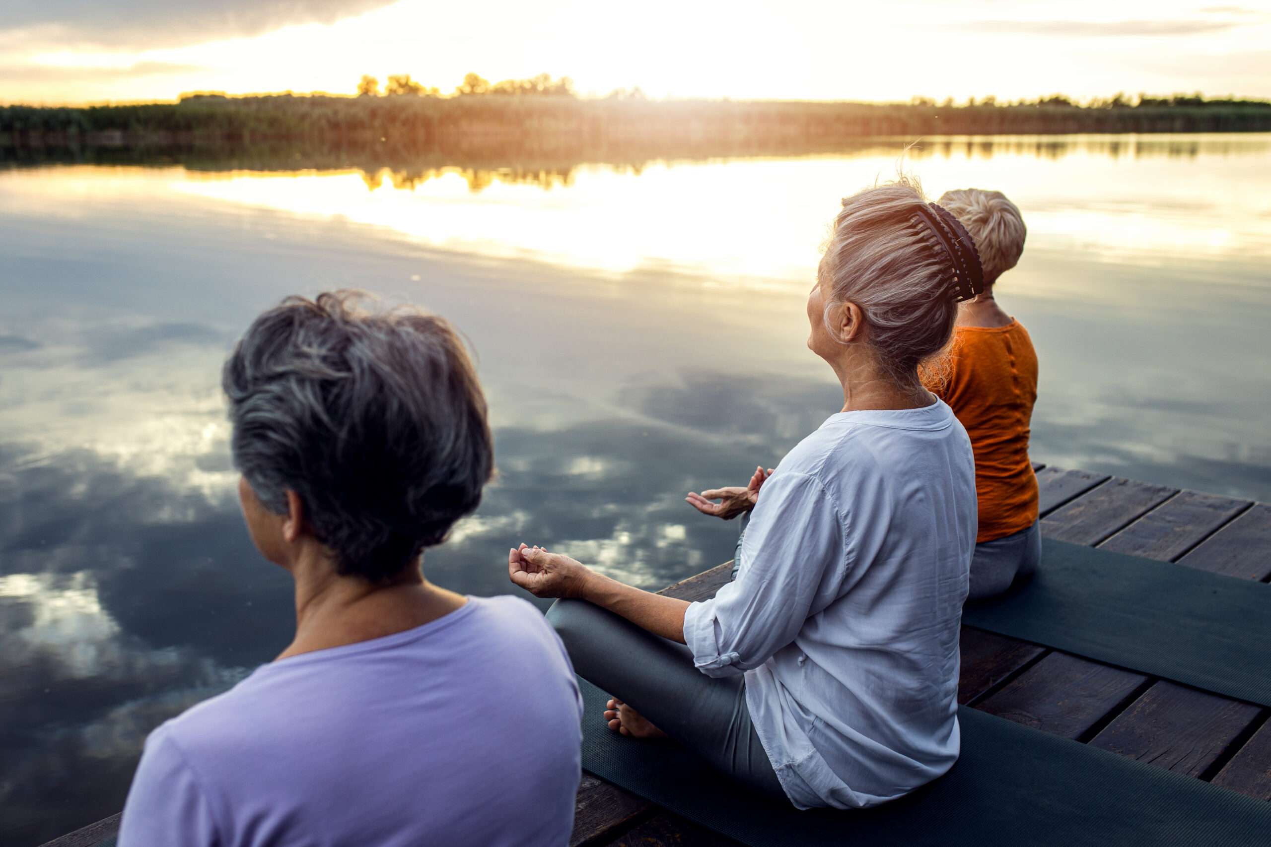 Three women sitting cross-legged on a lakeside dock at sunset during a daily meditation practice