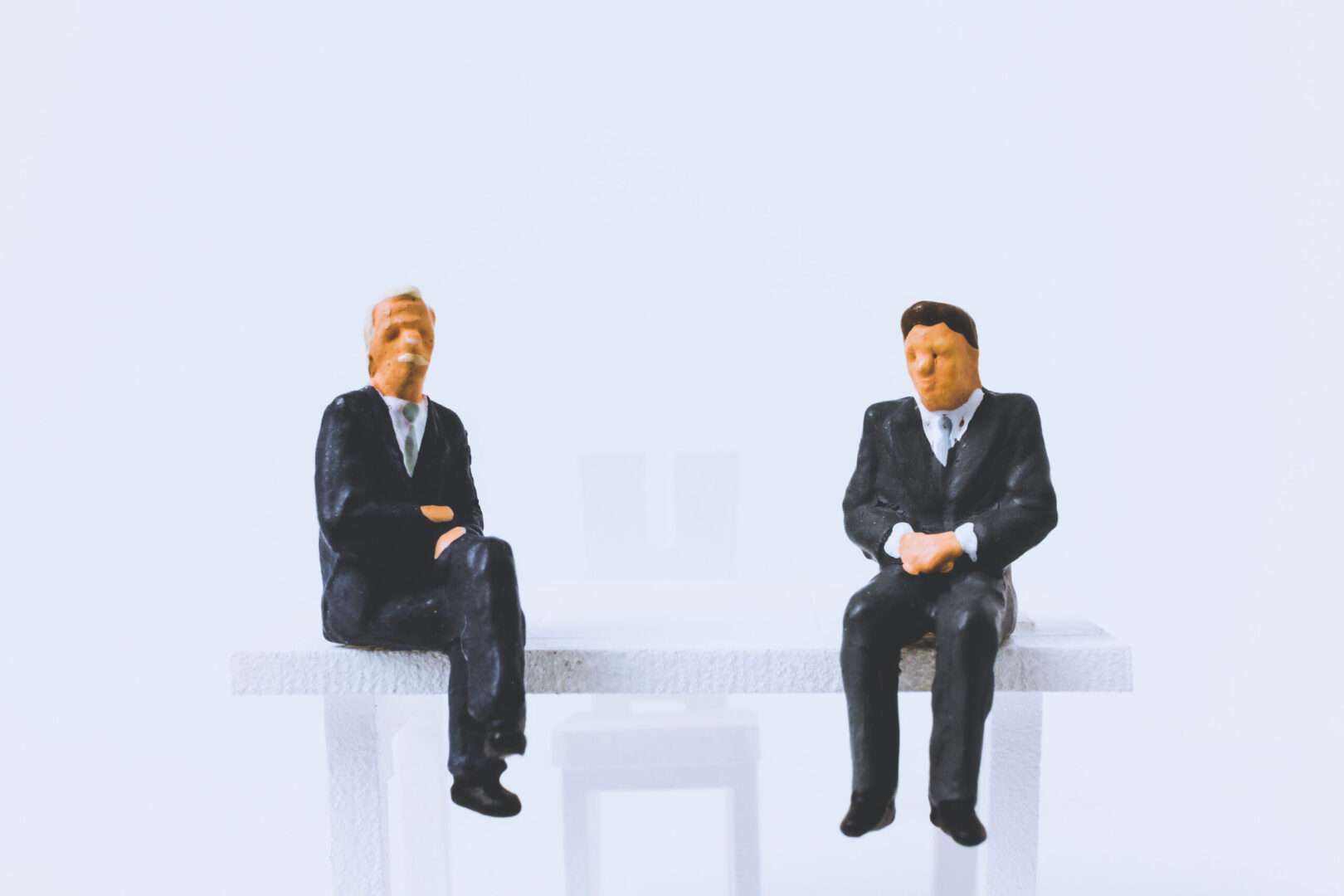 Two miniature figures in suits seated apart on a white bench, representing the difference between a board seat and an observer seat