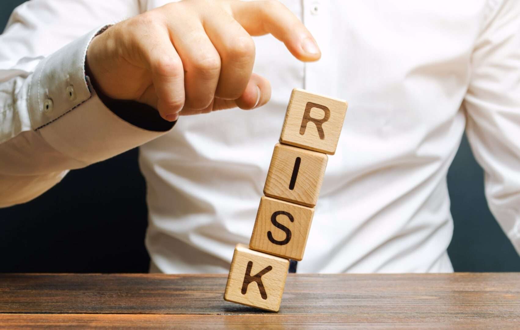 Hand about to flick a leaning stack of wooden blocks spelling RISK, representing how a corporate structure can collapse under liability